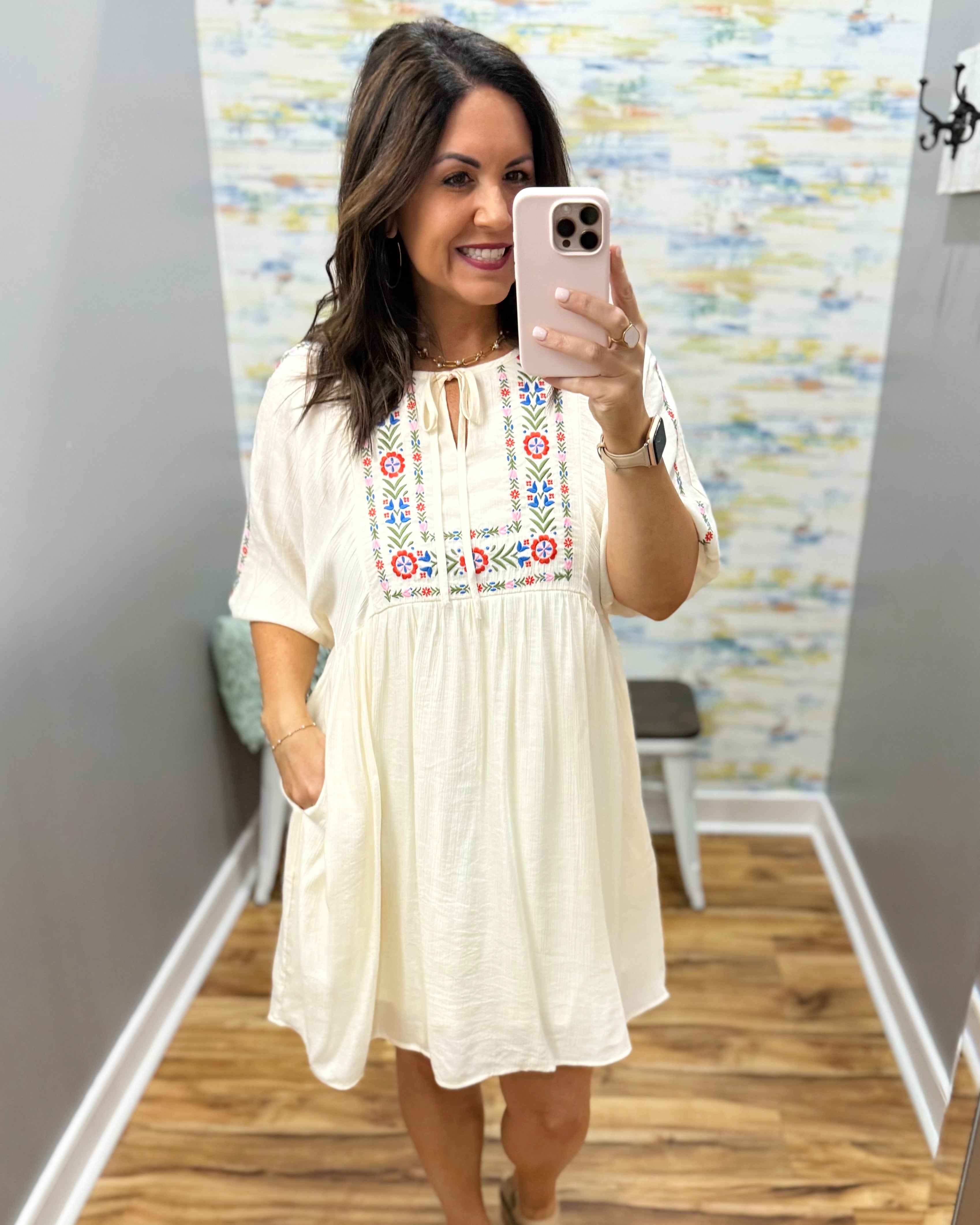 Woman taking a mirror selfie wearing a white dress with colorful embroidery in a room.