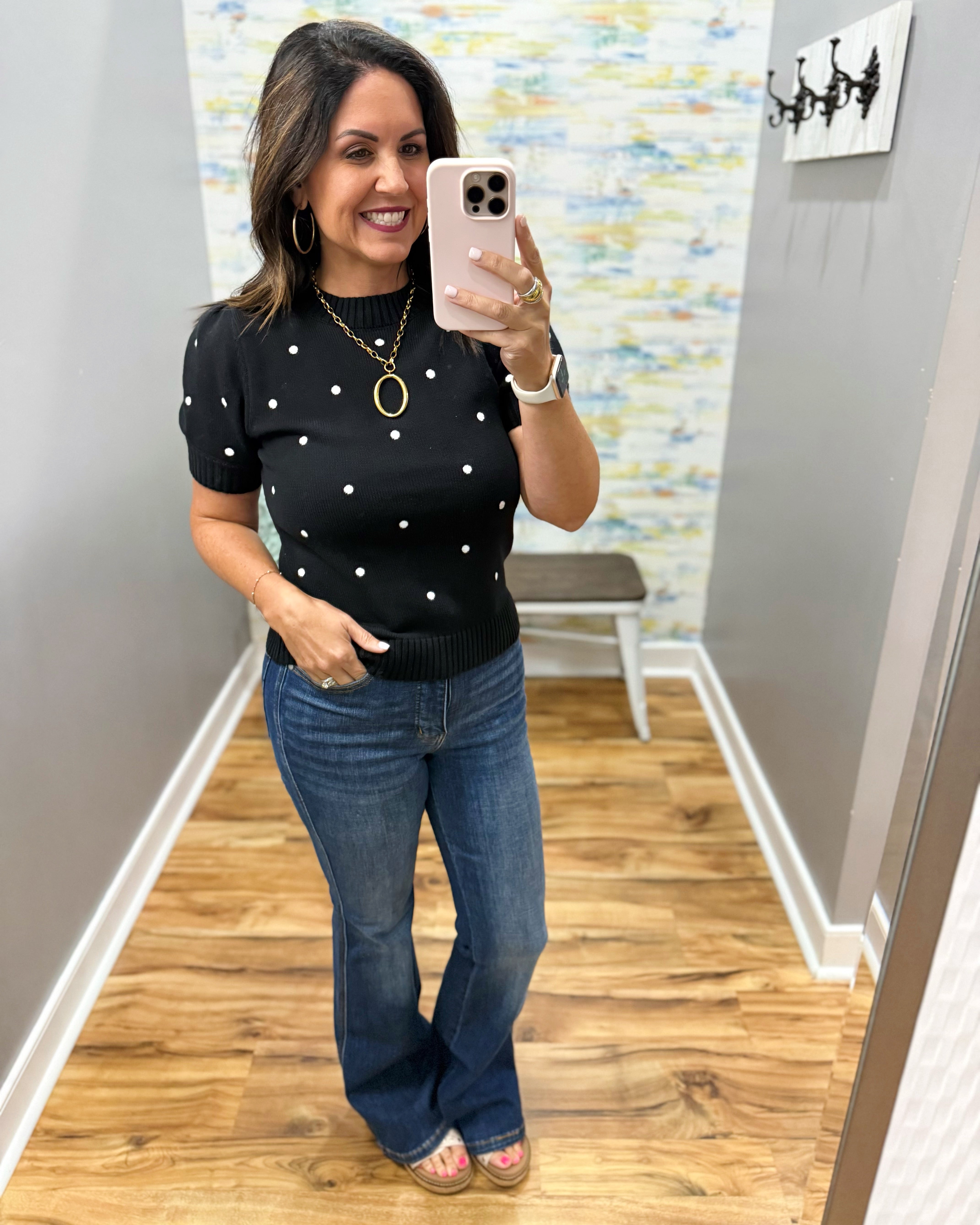 A woman takes a mirror selfie in a fitting room wearing a black short-sleeve knit top with white polka dots and blue jeans.
