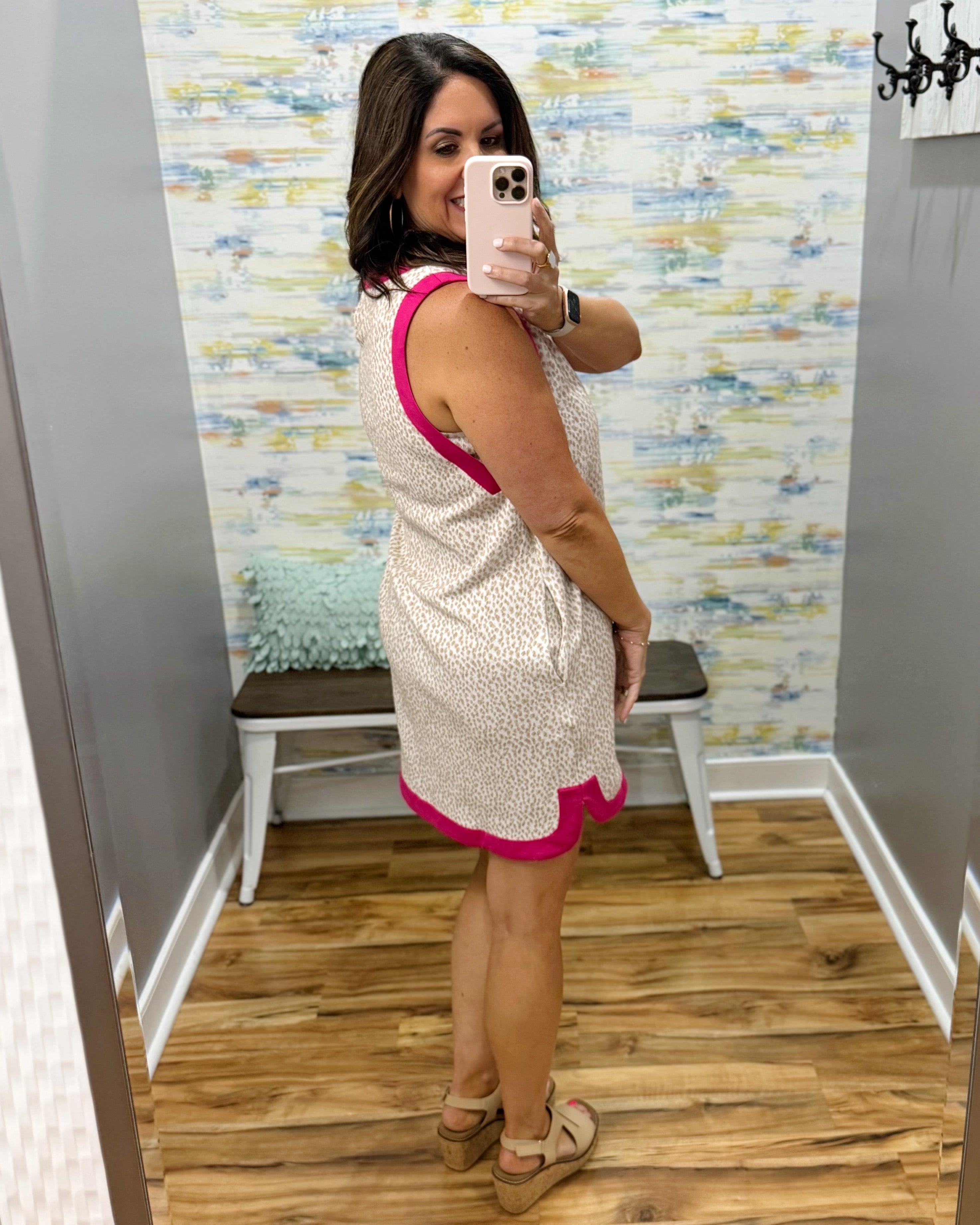 Woman taking a mirror selfie wearing a sleeveless dress with pink trim in a room with patterned wallpaper.