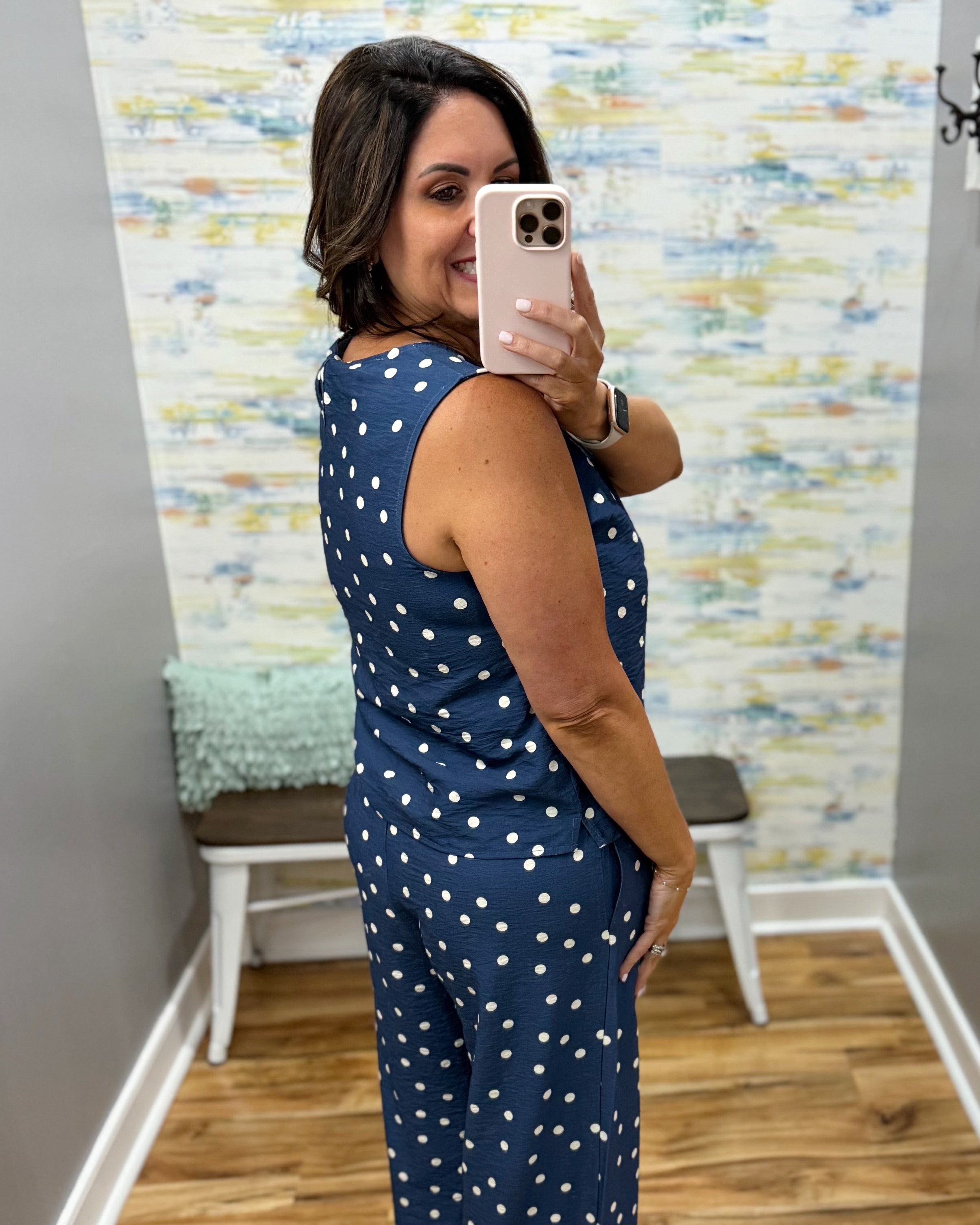 Woman taking a mirror selfie wearing a blue polka dot jumpsuit in a room with a textured wall.