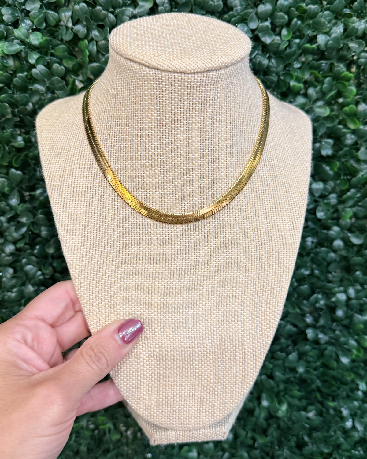 Chansuttpearls Herringbone gold snake chain necklace displayed on a beige fabric jewelry bust, held by a hand against a green leafy hedge background.