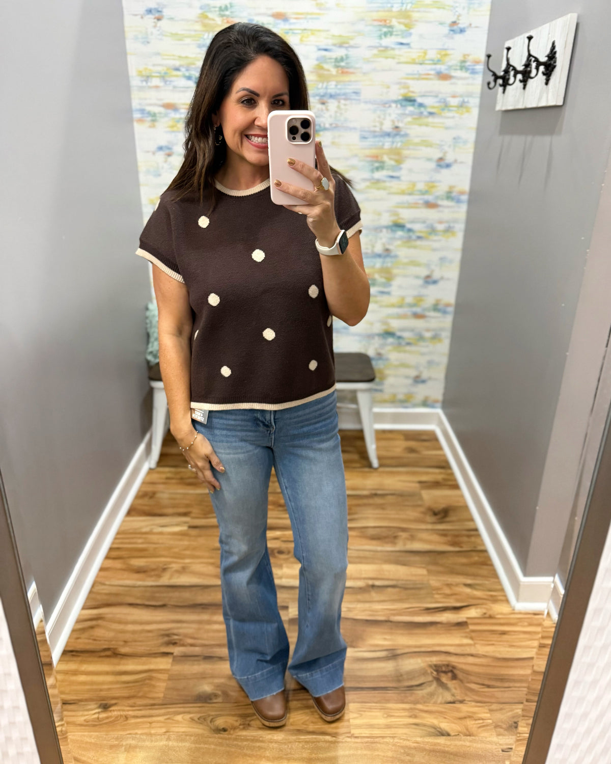 A woman takes a mirror selfie wearing a brown short-sleeve knit top with cream trim and cream polka dots, paired with blue jeans and brown shoes, in a fitting room.
