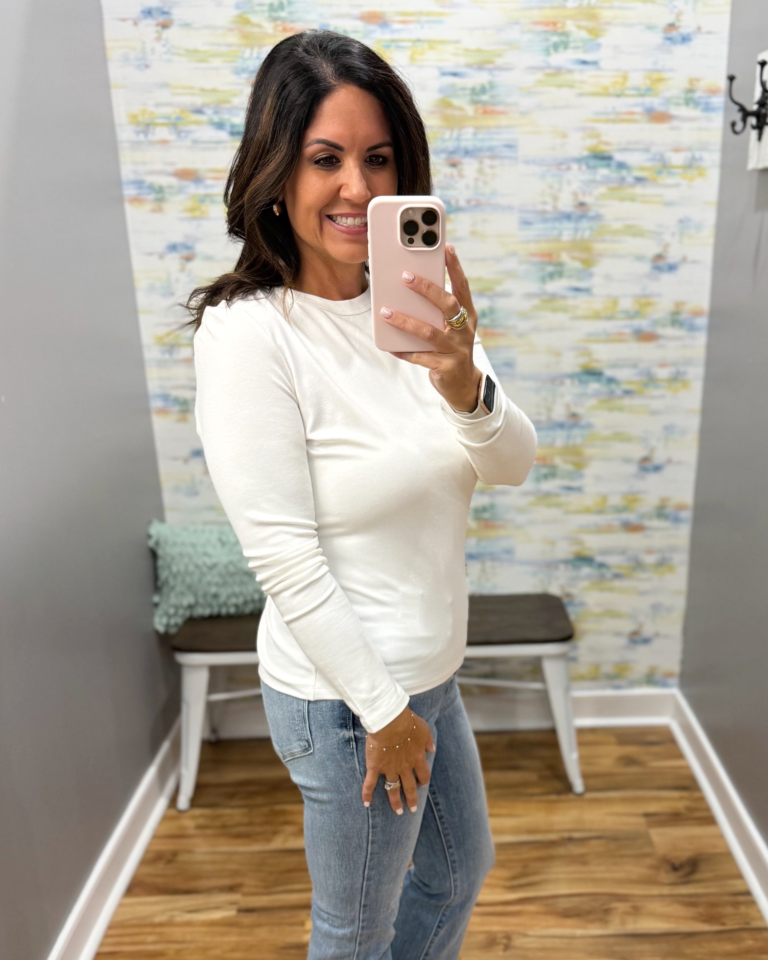 A woman taking a mirror selfie in a fitting room wearing a fitted long-sleeve white top and light blue jeans.