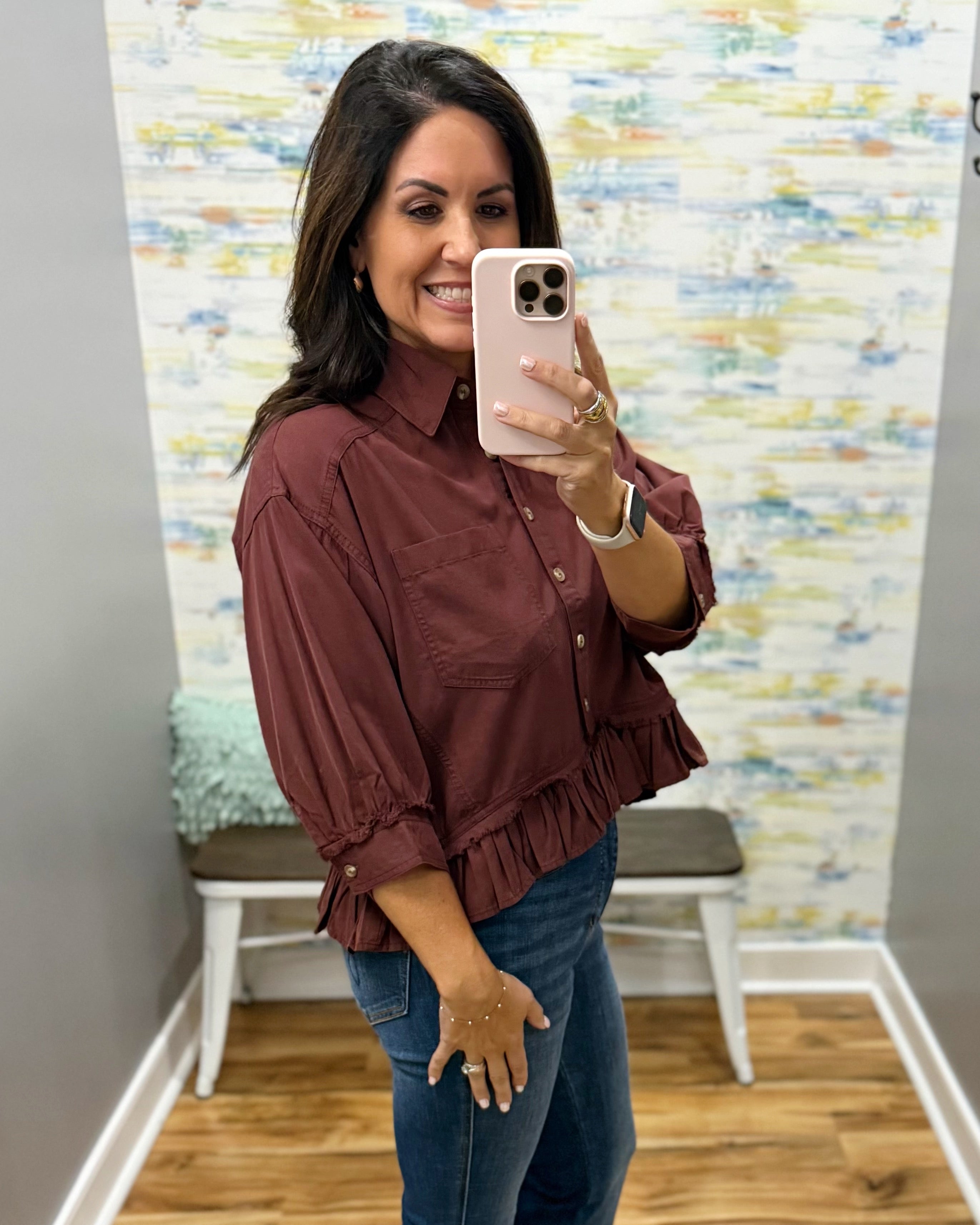 A woman takes a mirror selfie in a boutique showing a rust-brown ruffled blouse with three-quarter sleeves and a button-down front, paired with blue jeans.