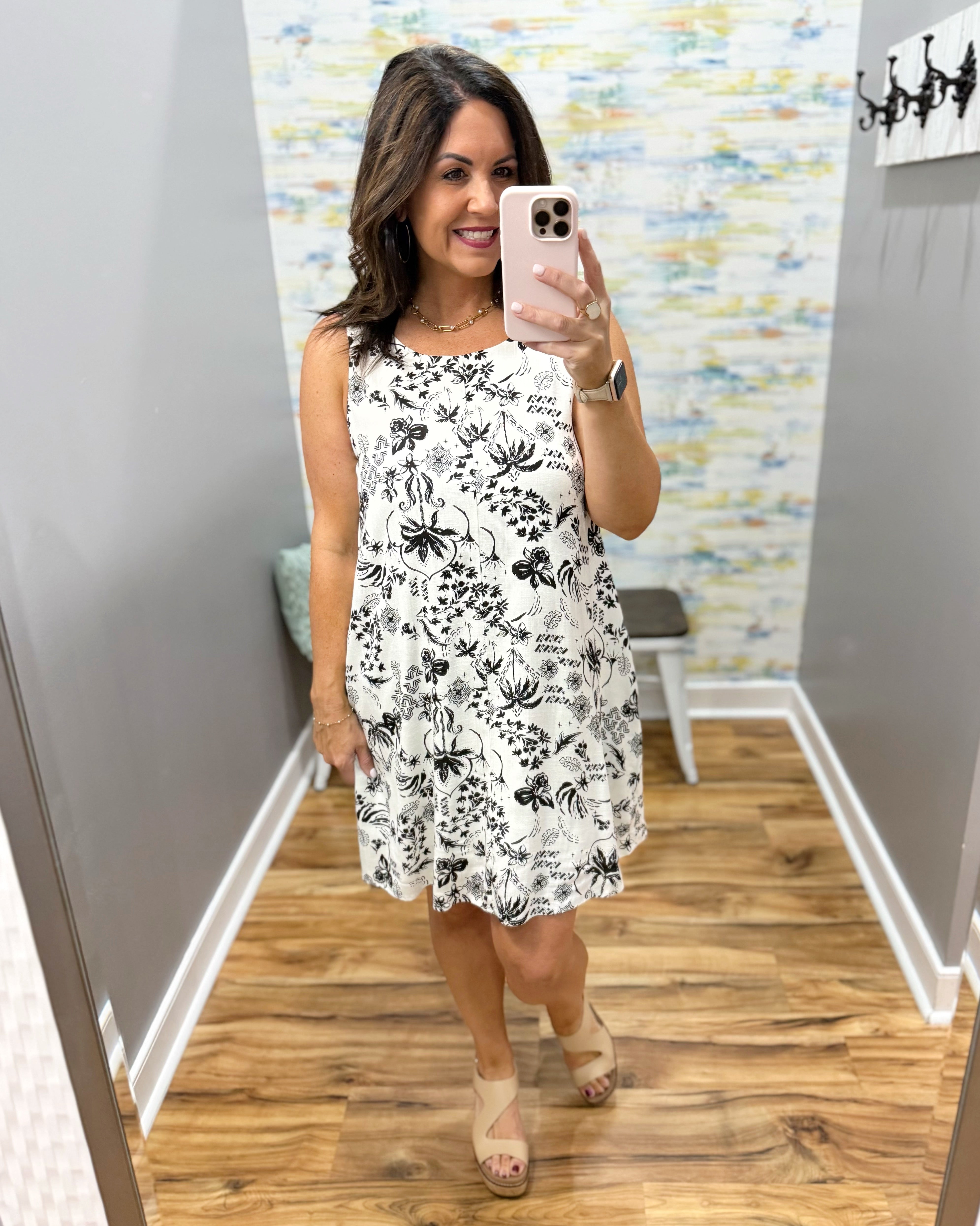 Woman taking a mirror selfie wearing a white dress with black floral pattern in a room with a colorful wall.