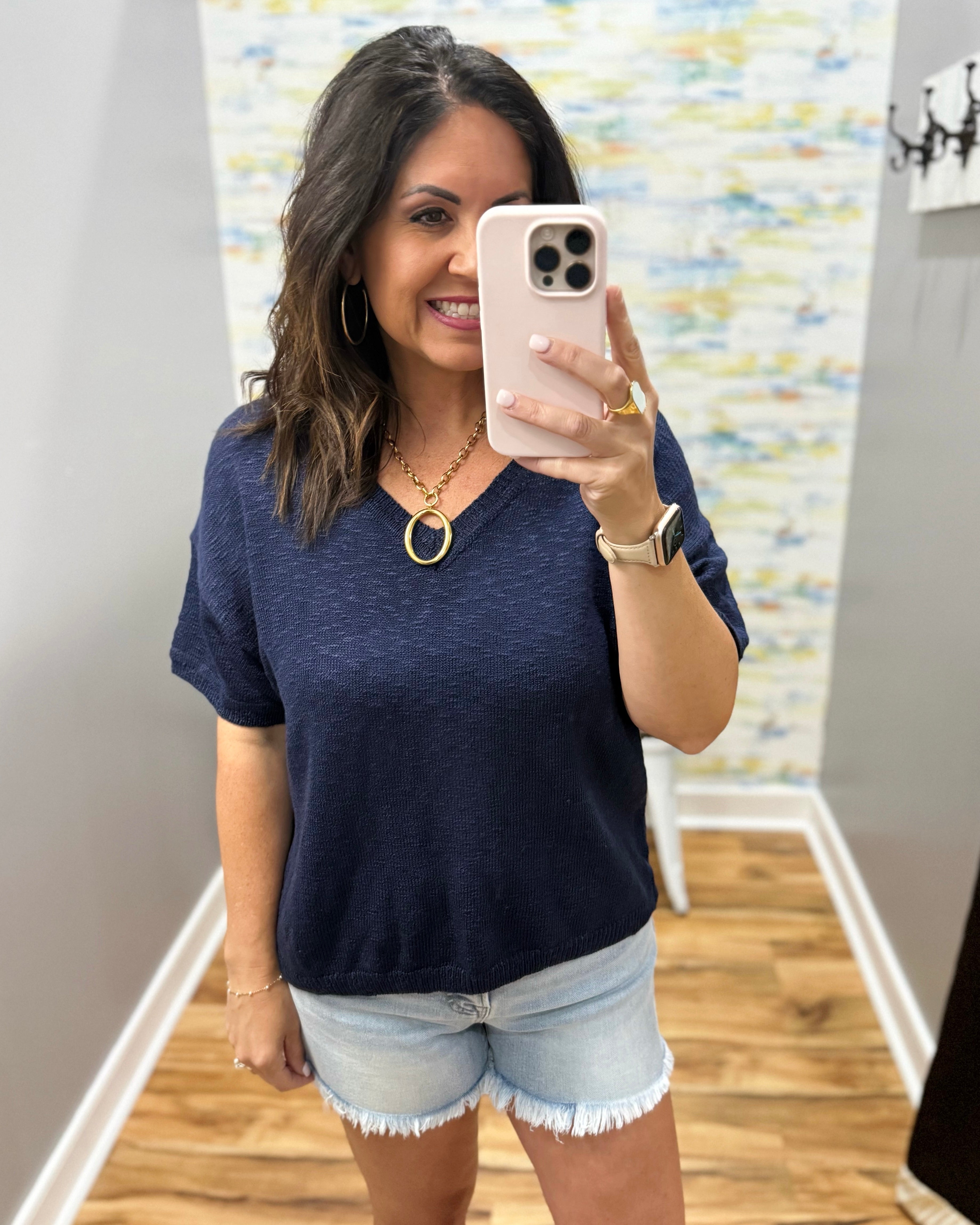 Woman taking a mirror selfie wearing a navy blue t-shirt and light denim shorts.