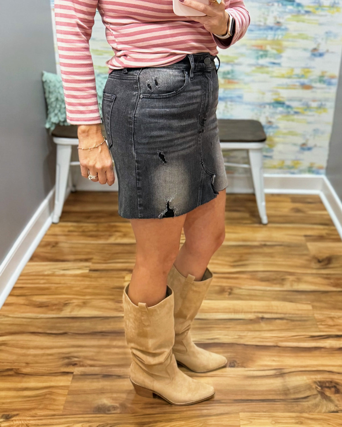 A person wearing a distressed black denim mini skirt, a pink and white striped long-sleeve top, and beige suede knee-high boots, standing on a wood floor in an indoor room.