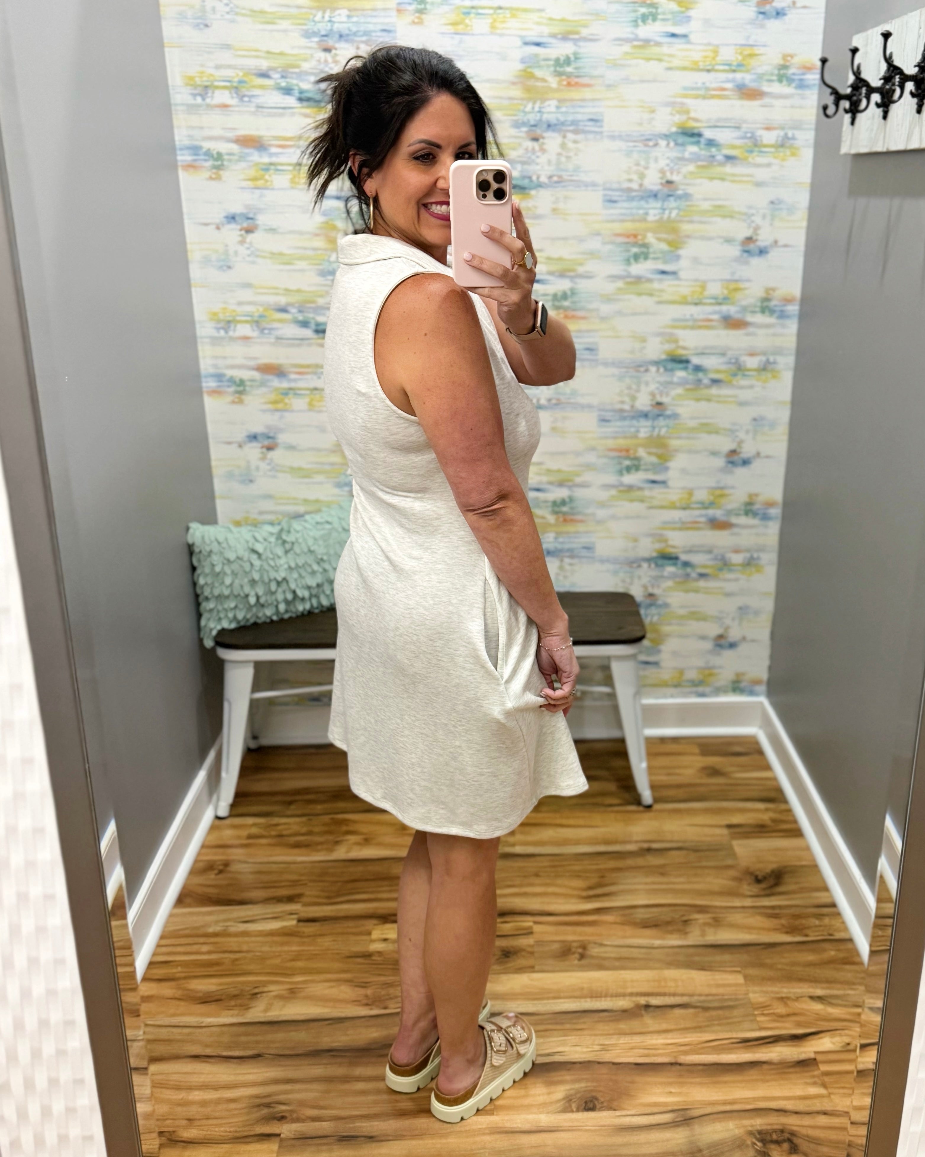Woman taking a mirror selfie wearing a white dress in a room with patterned wallpaper.