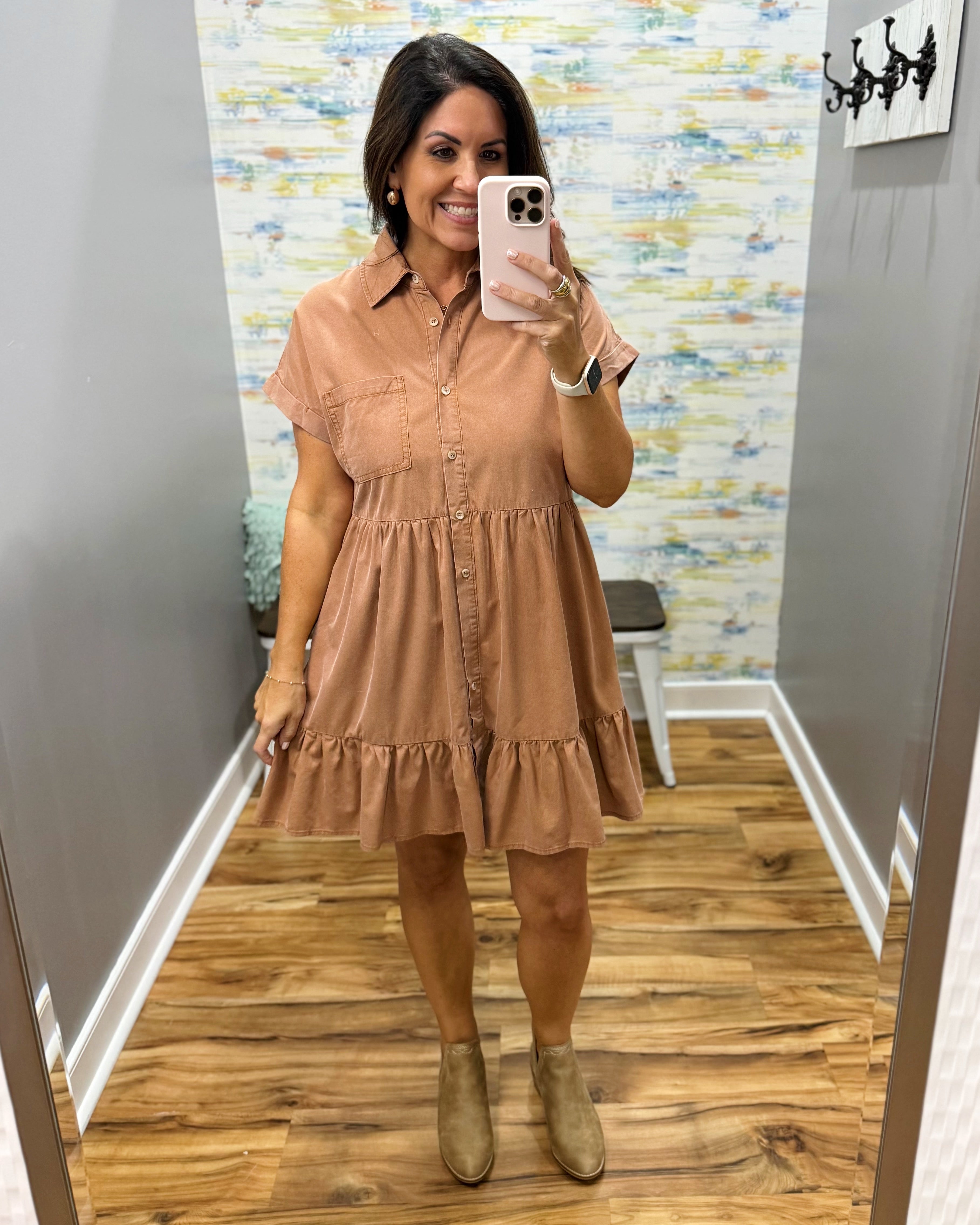 A woman takes a mirror selfie in a fitting room wearing a short-sleeve button-front shift dress in a warm tan/camel color, paired with tan ankle boots.