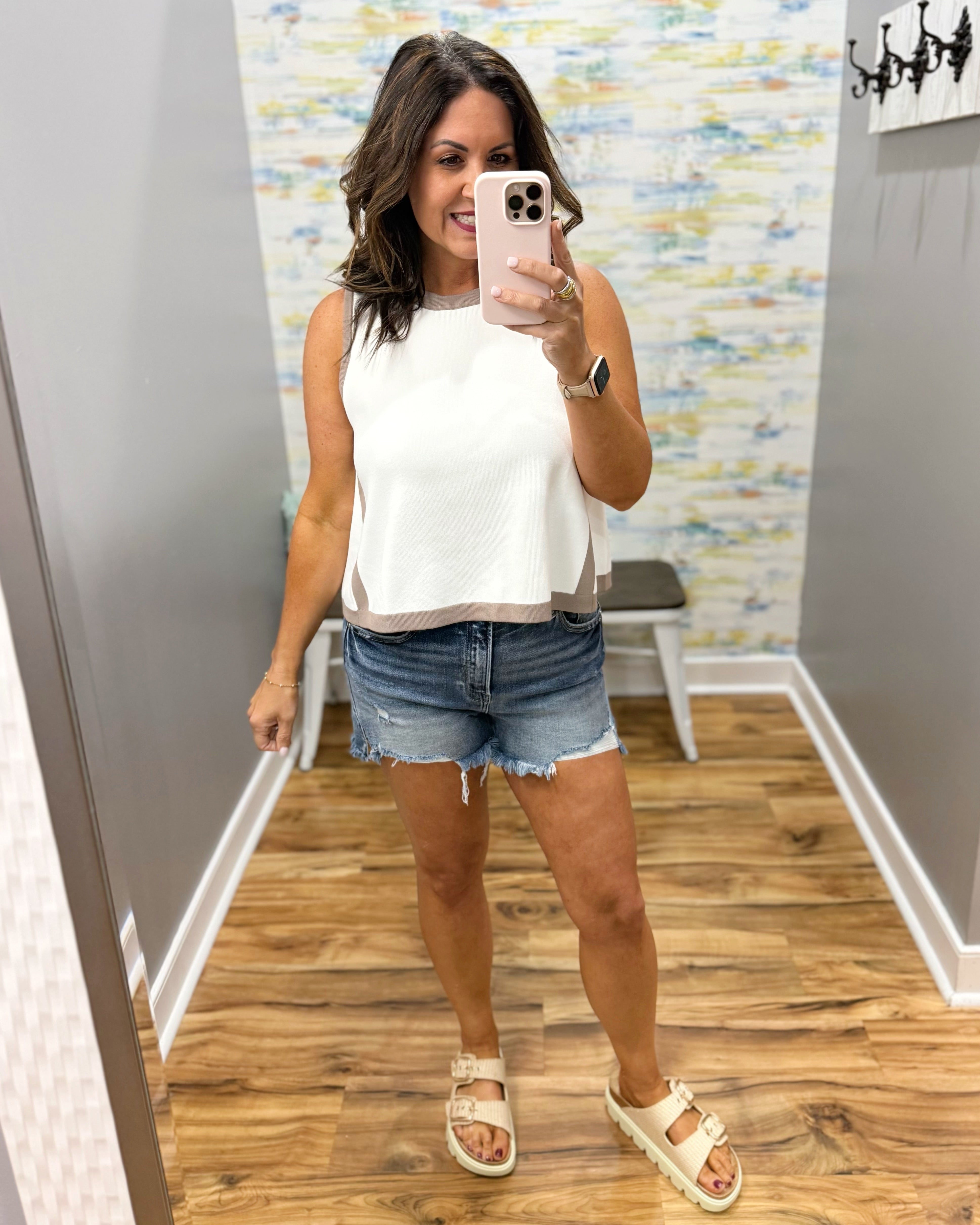 Woman taking a mirror selfie wearing a white sleeveless top and denim shorts in a room with a colorful wall.