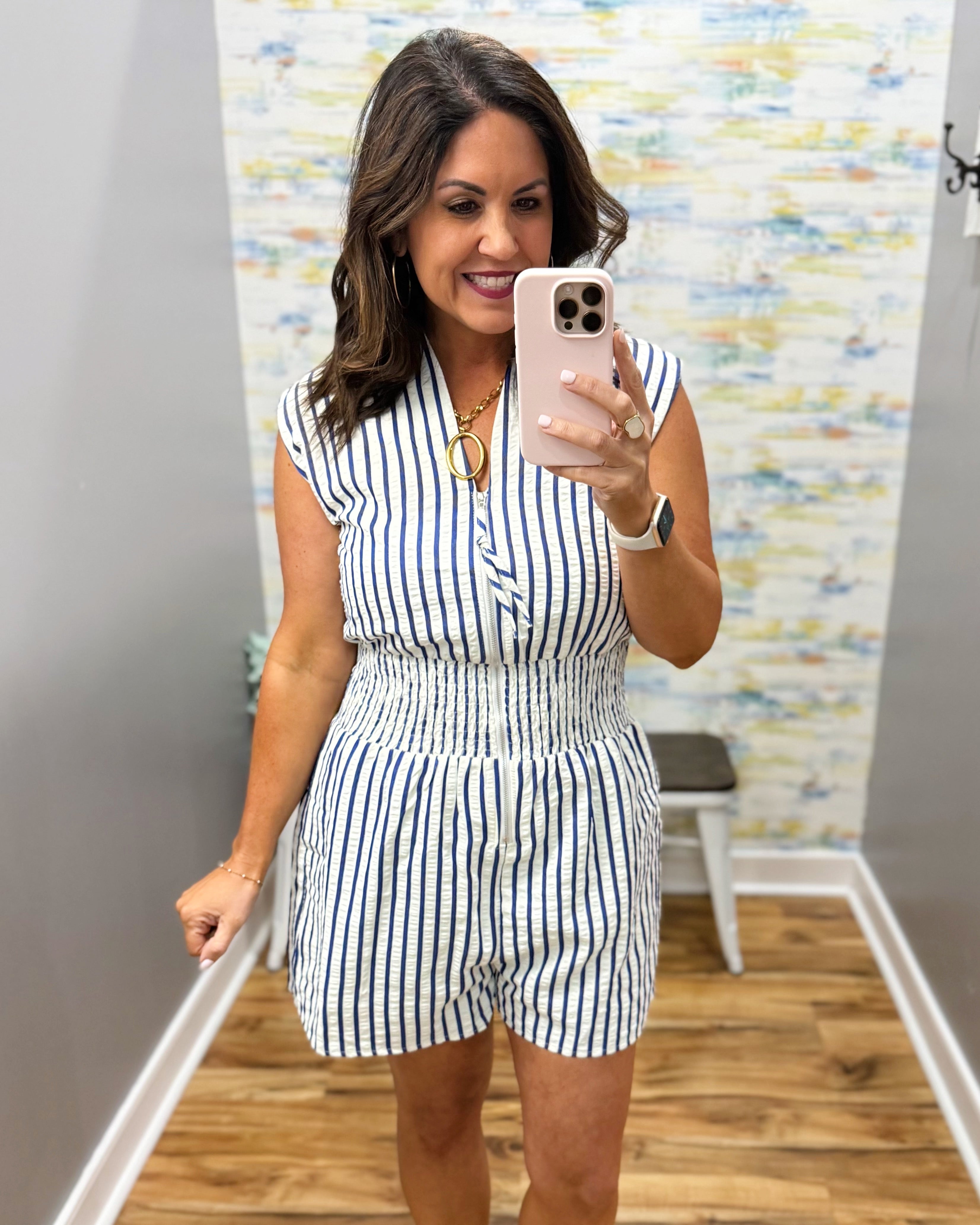 Woman taking a mirror selfie wearing a striped romper in a room with a colorful wall.