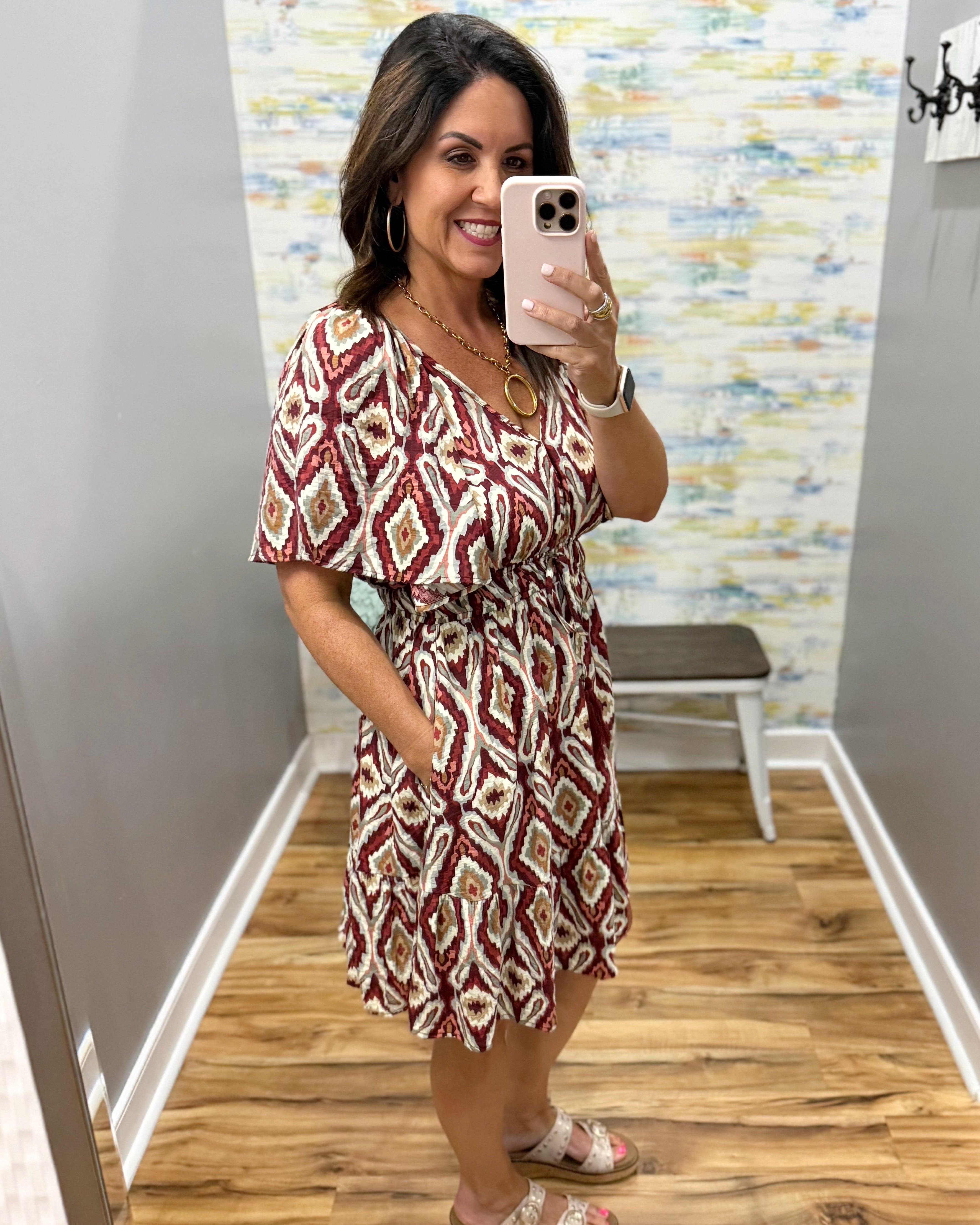 A woman takes a mirror selfie in a boutique wearing a short-sleeve V-neck dress with a red, beige, and white geometric diamond pattern. The background shows a gray wall, a colorful abstract wallpaper, a wooden floor, and a small bench.