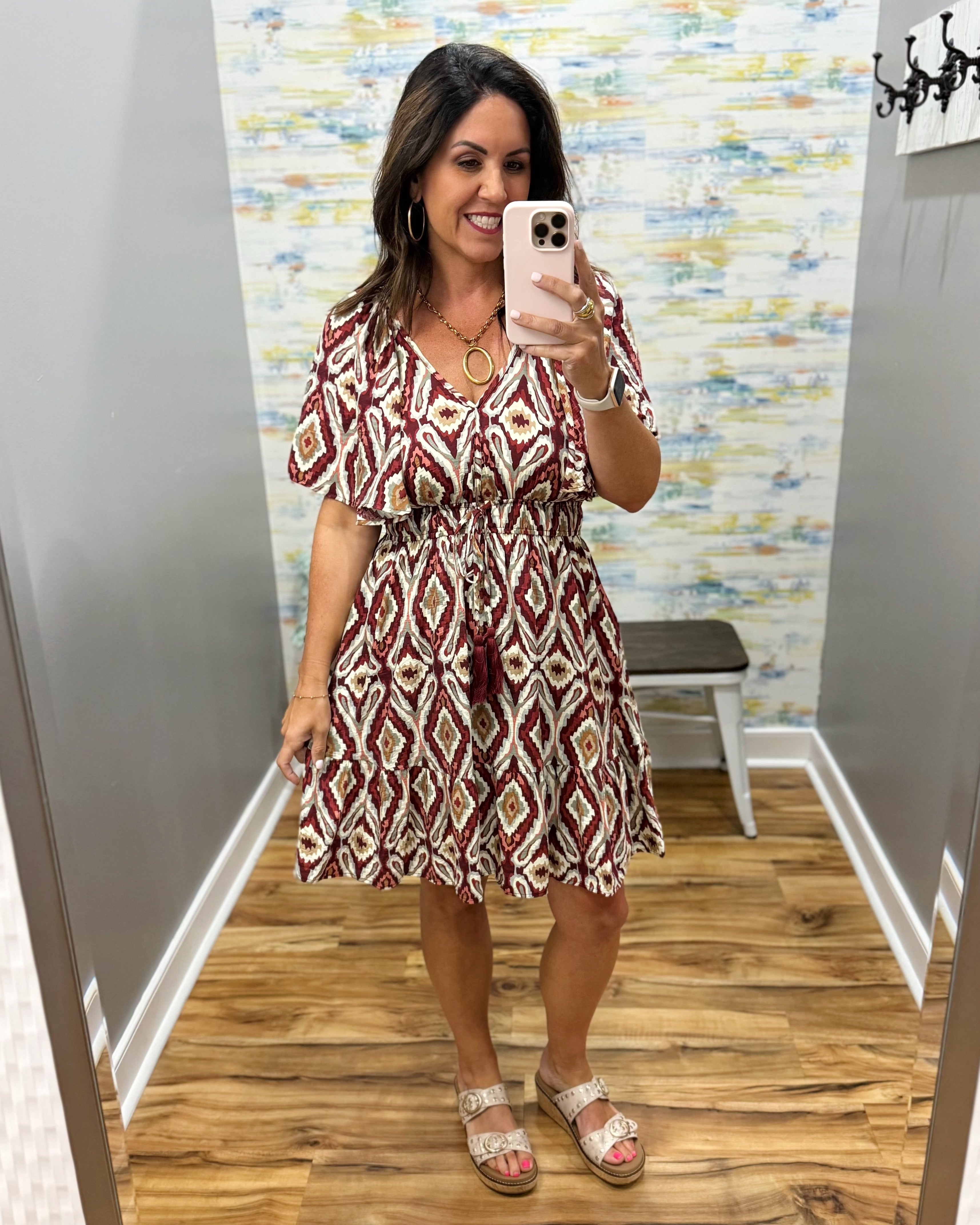 A woman takes a mirror selfie in a fitting room wearing a short-sleeve V-neck dress with a red, beige, and white geometric diamond pattern. She is wearing beige sandals and holding a light pink smartphone.