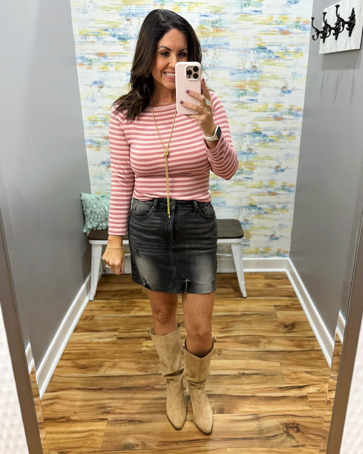 A woman takes a mirror selfie in a fitting room wearing a ginger and mauve striped long-sleeve top, a dark gray denim mini skirt, and beige knee-high boots.