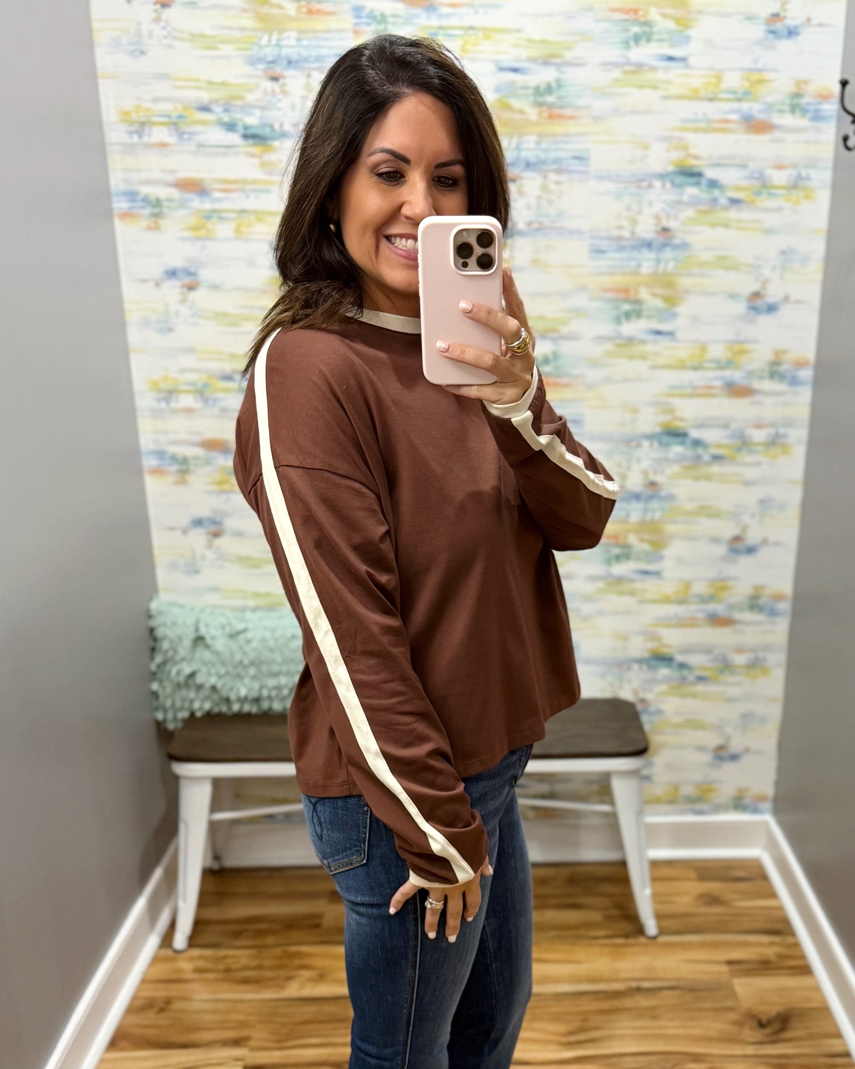 A woman takes a mirror selfie wearing a brown long-sleeve top with cream-colored stripes down the sleeves and a matching cream crew neck. She is wearing blue jeans and holding a light pink smartphone.