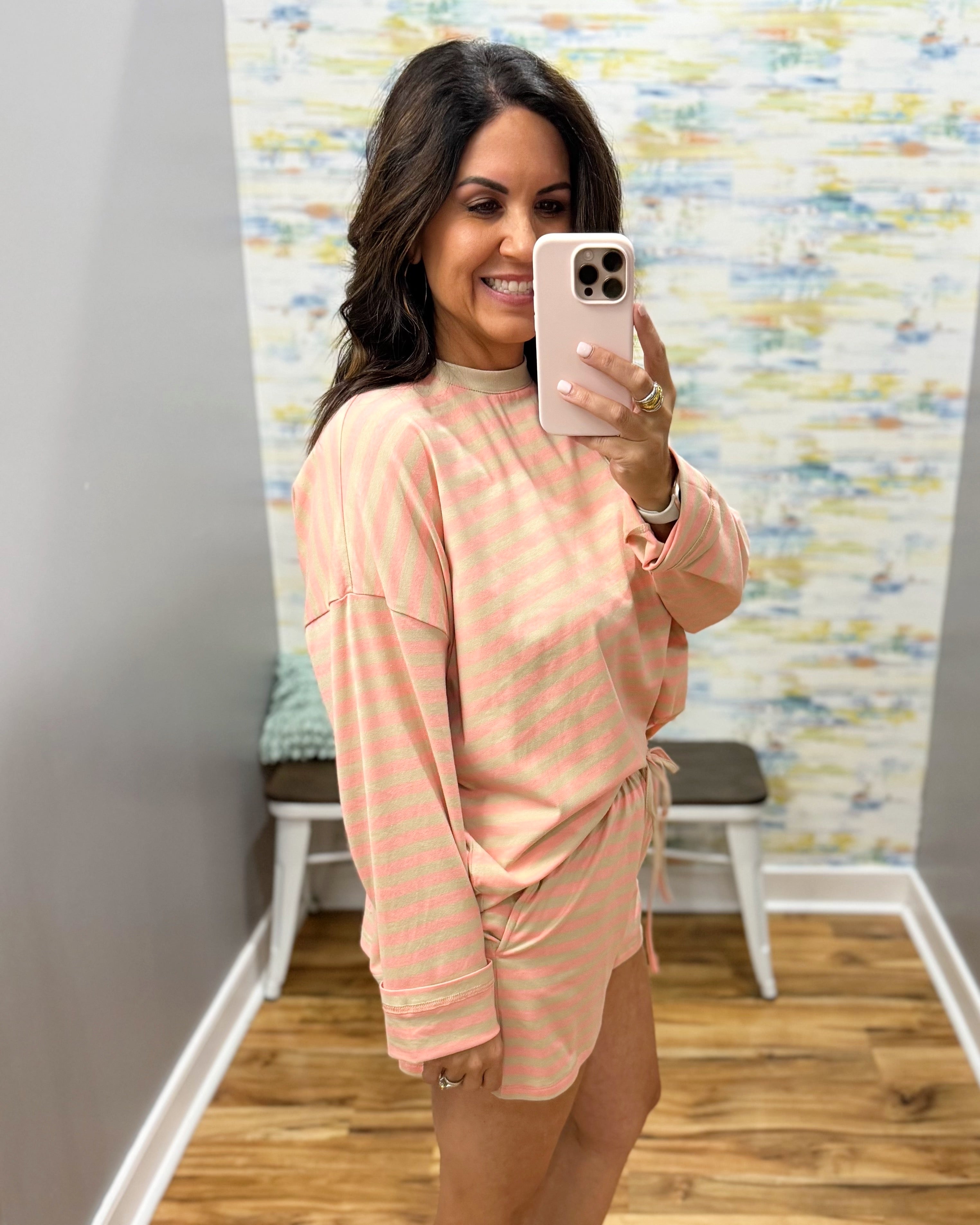 A woman takes a mirror selfie wearing a light peach and white striped long-sleeve tunic top with a matching tie-waist skirt. The setting is a home interior with a wood floor and a patterned wallpaper background.