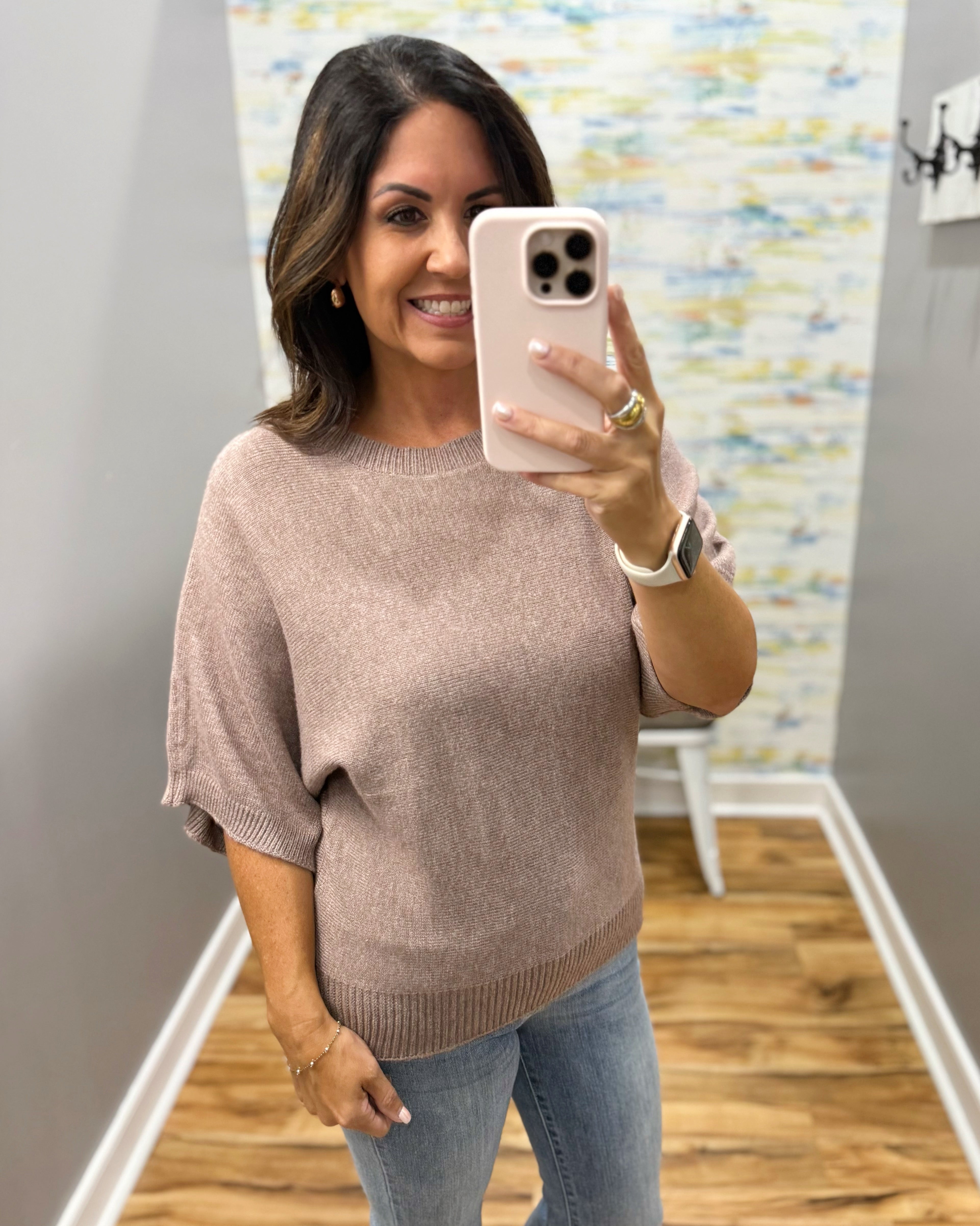 A woman takes a mirror selfie wearing a light taupe knit sweater with elbow-length sleeves and light blue jeans in a home setting.