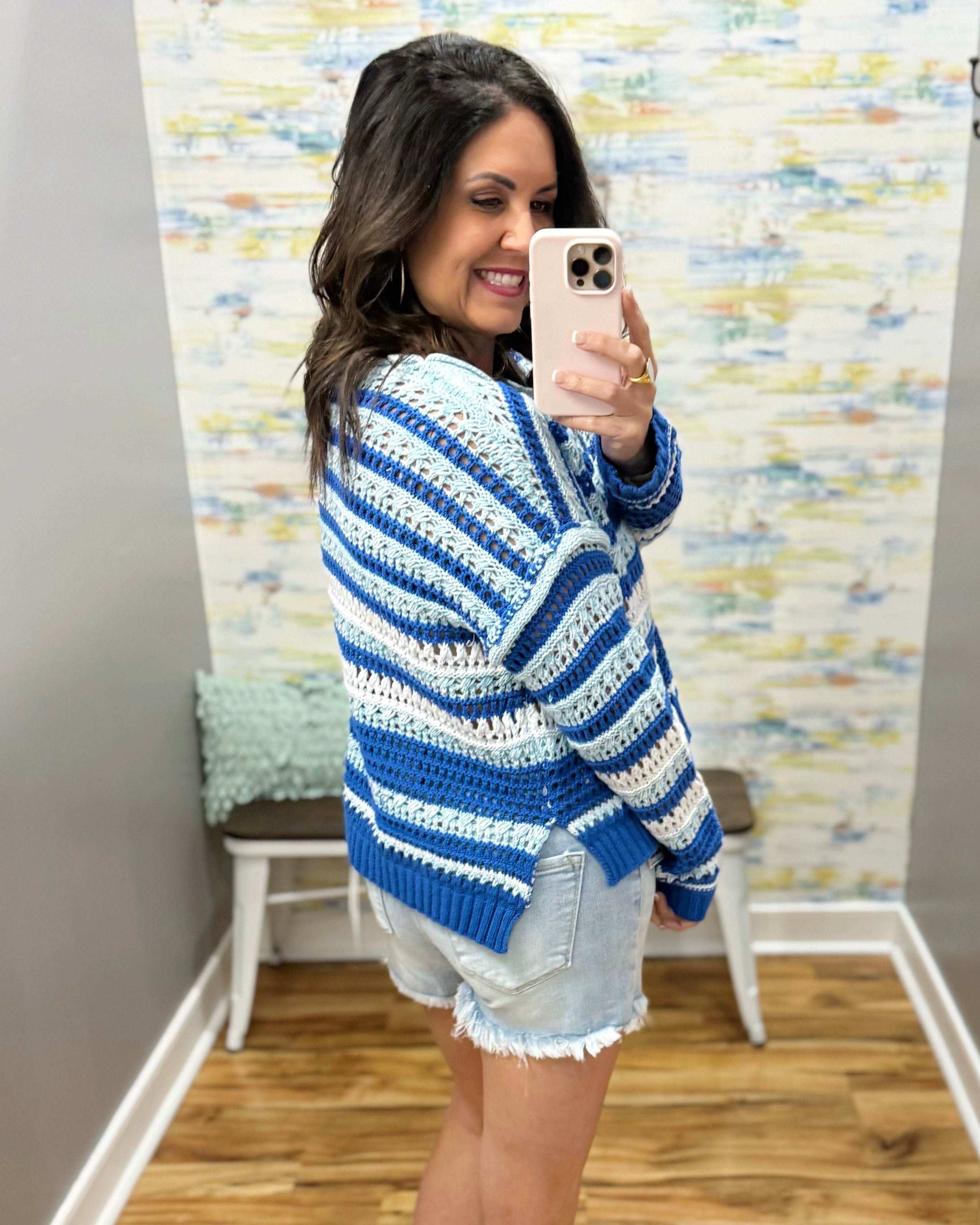Woman taking a mirror selfie wearing a blue and white striped sweater and denim shorts.