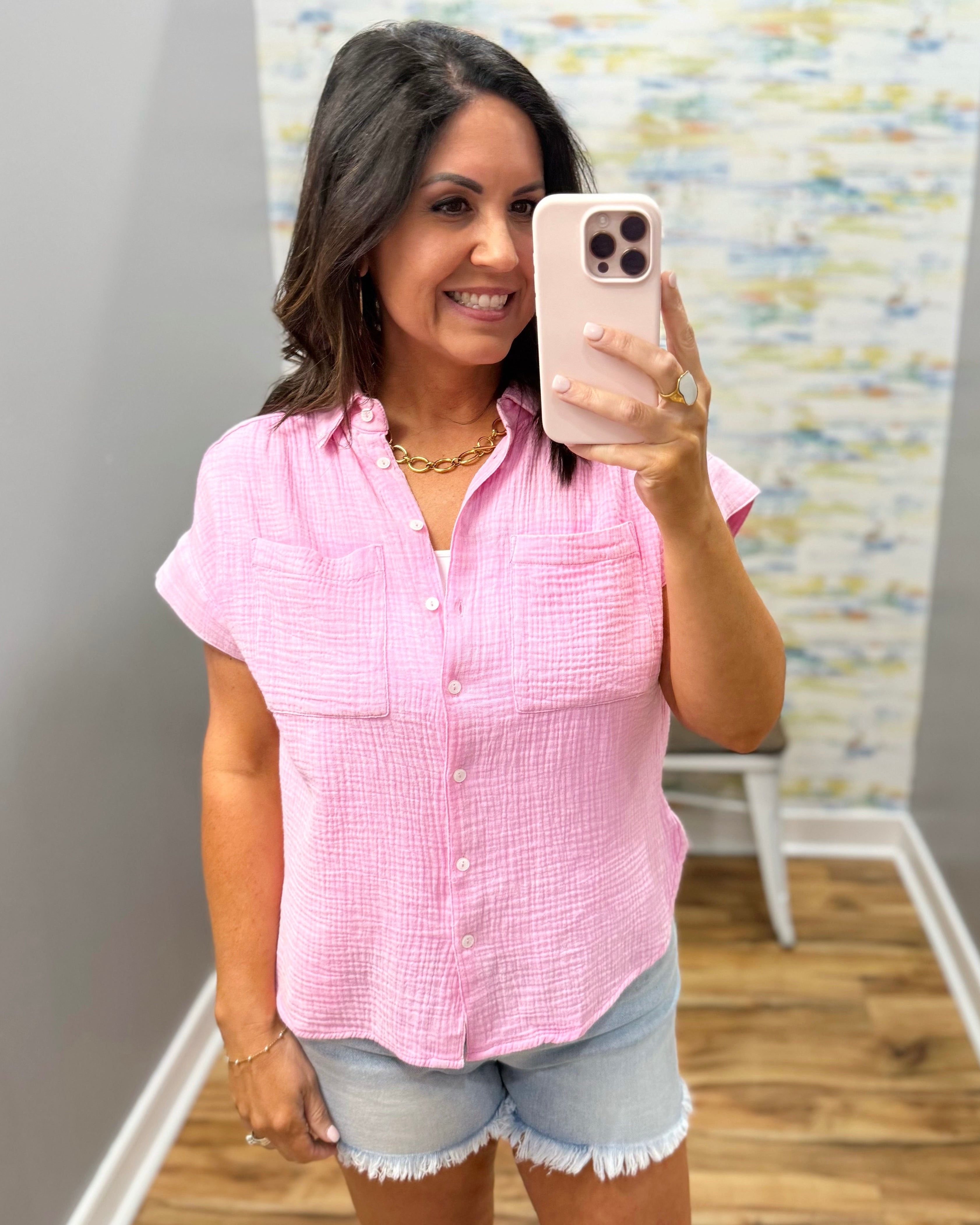 Woman taking a mirror selfie wearing a pink checkered shirt and denim shorts.