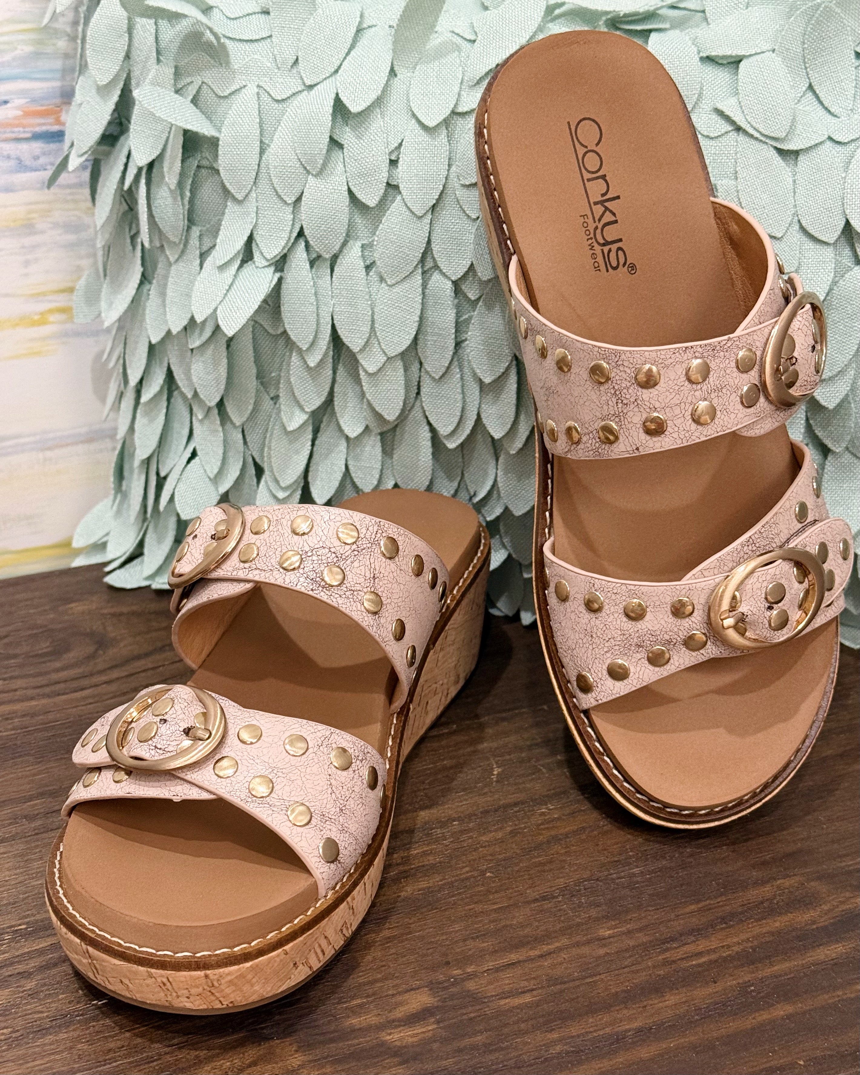 Pair of pink sandals with gold accents on a wooden surface with a decorative green leafy background.