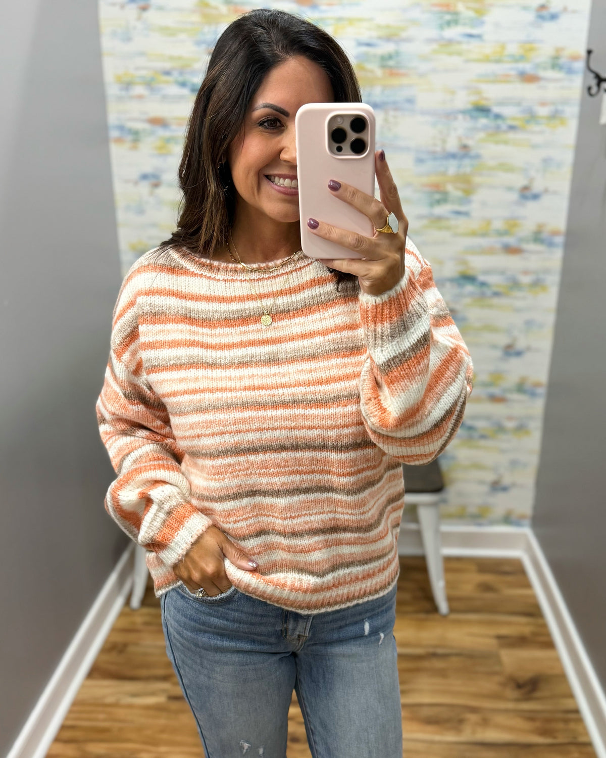 A woman taking a mirror selfie wearing a fuzzy, striped crewneck sweater in orange, beige, and white, paired with blue jeans.