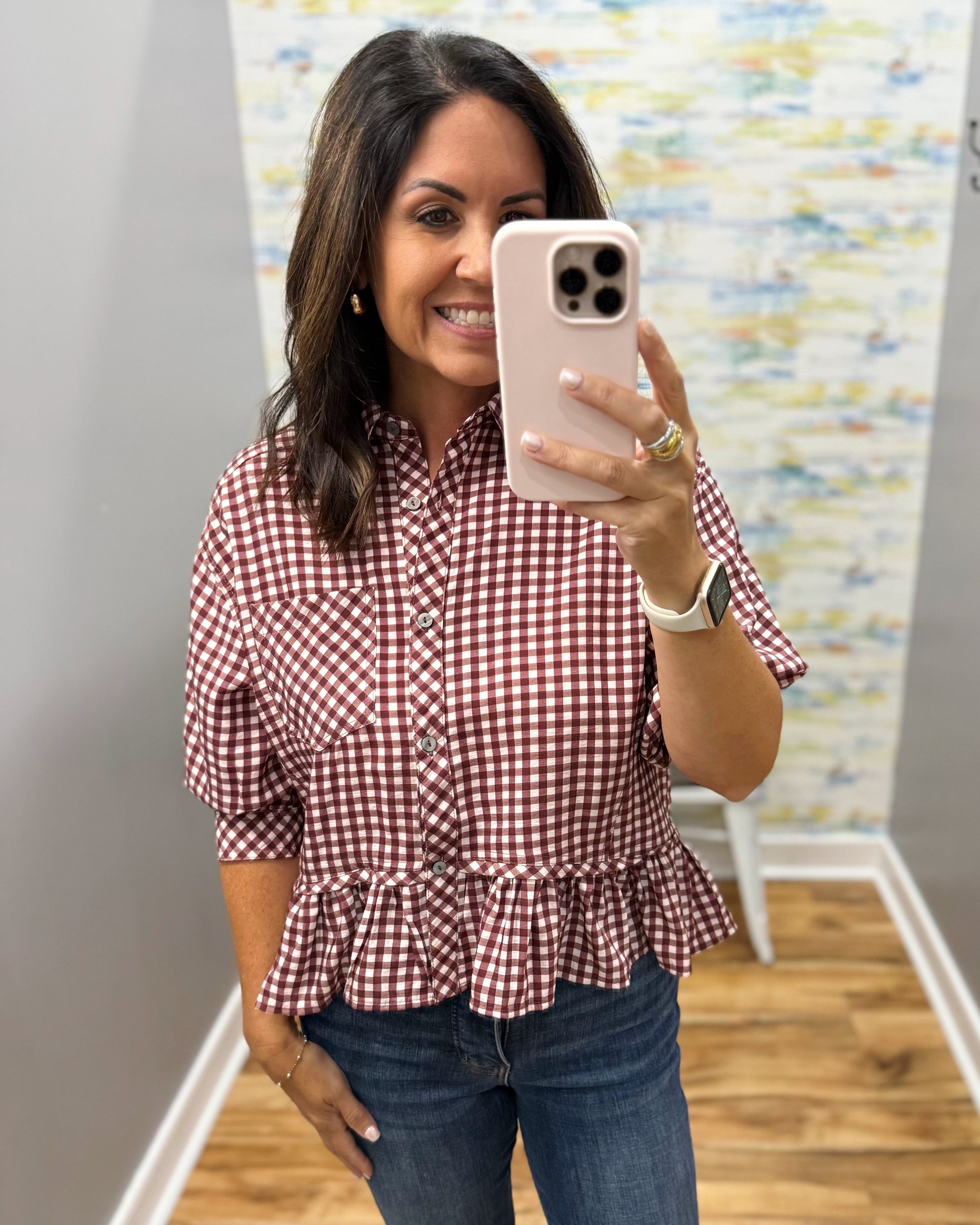 A woman taking a mirror selfie wearing a red and white gingham button-down shirt with puff sleeves and a ruffled hem, paired with blue jeans.