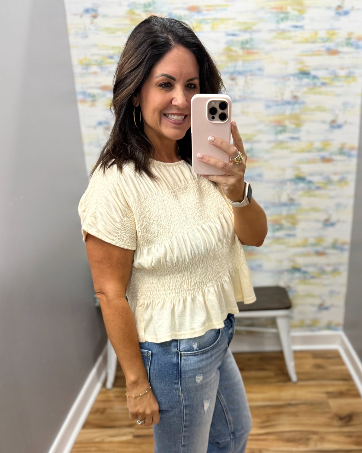 A woman takes a mirror selfie wearing a cream-colored, short-sleeved textured top and blue jeans in a fitting room.
