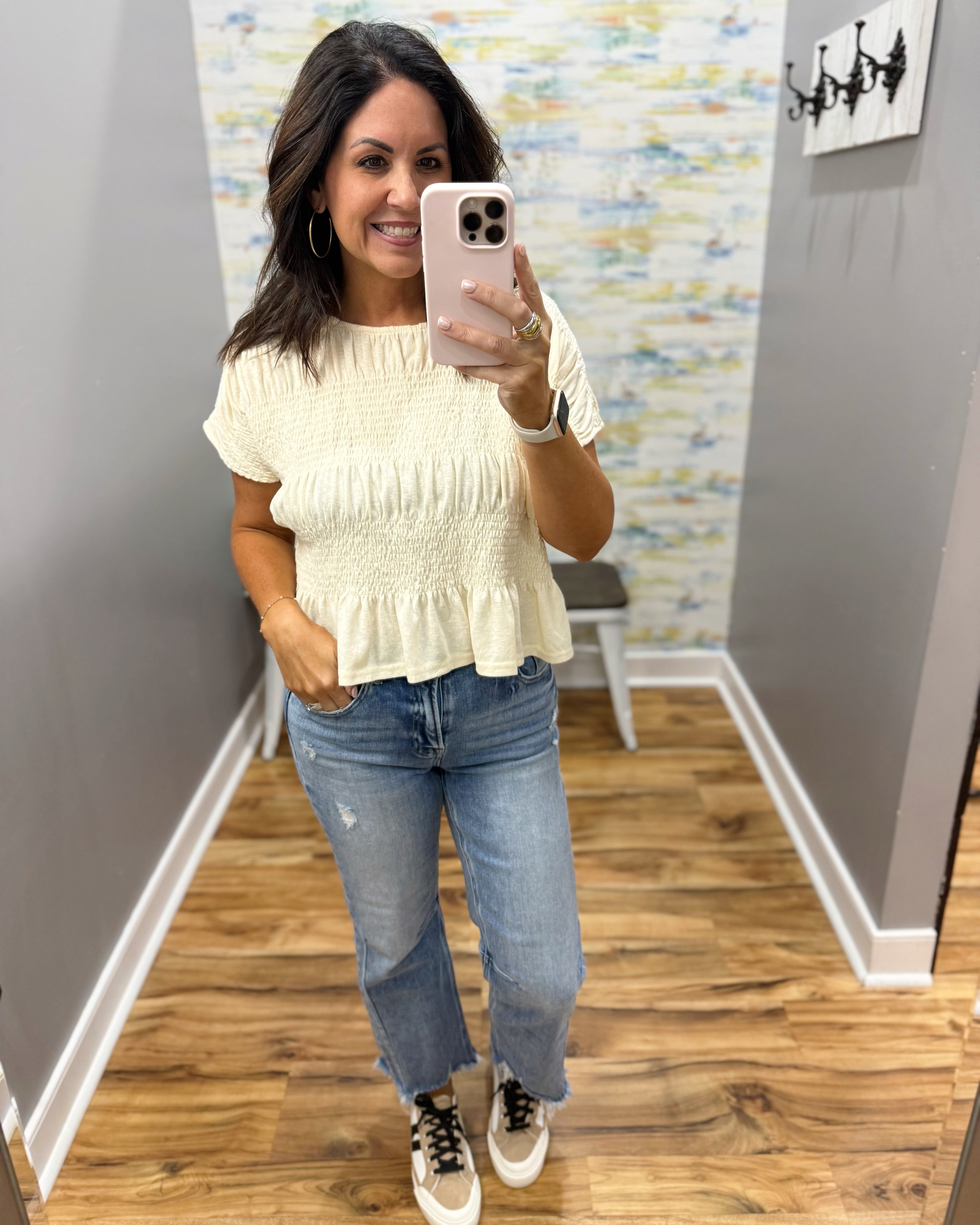 A woman takes a mirror selfie in a fitting room wearing a cream-colored textured short-sleeve ruffle top, blue jeans, and white sneakers with black laces.