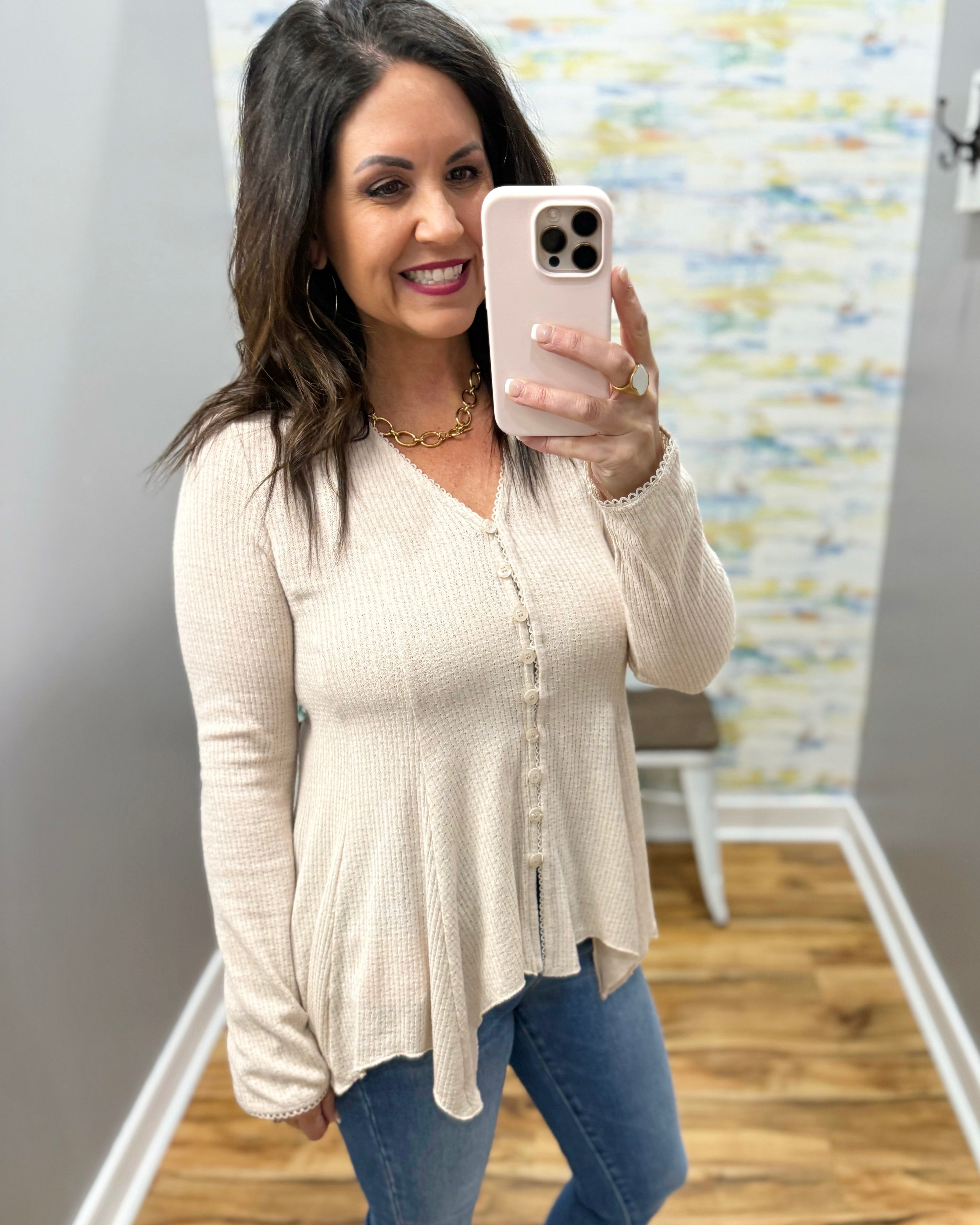 Woman taking a mirror selfie wearing a beige cardigan and blue jeans in a room with a colorful wall.