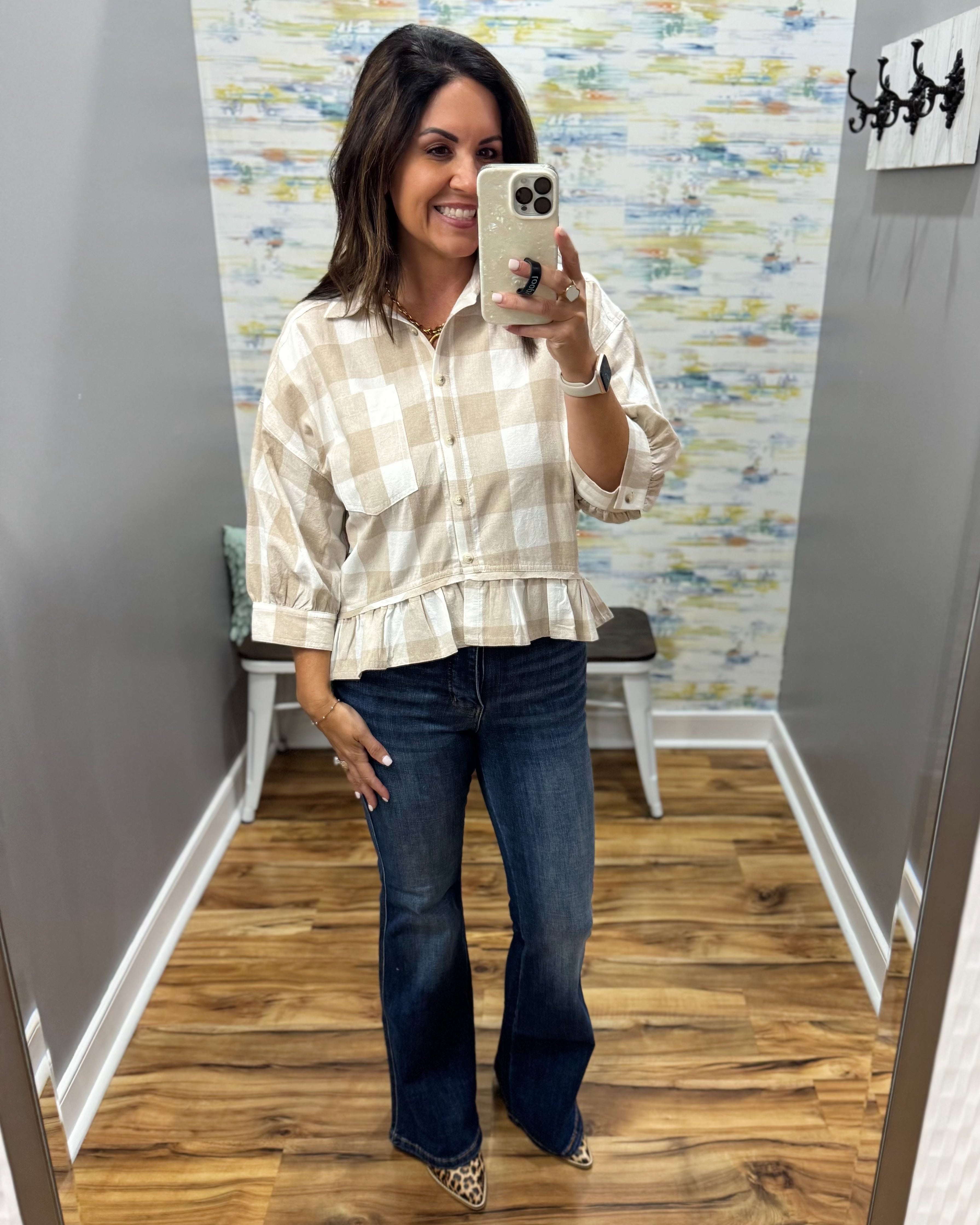 A woman takes a mirror selfie in a boutique showing a beige and white plaid button-down shirt with a ruffled hem and dark blue flare jeans.