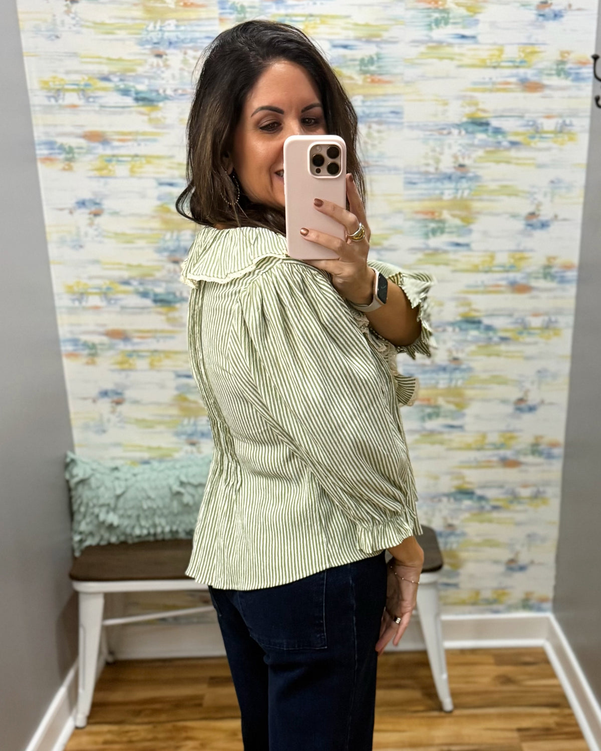 A woman takes a mirror selfie wearing a light sage green and white vertically striped blouse with ruffled shoulders and dark blue jeans. The background features a colorful abstract-patterned wallpaper and a small white bench with a mint green pillow.