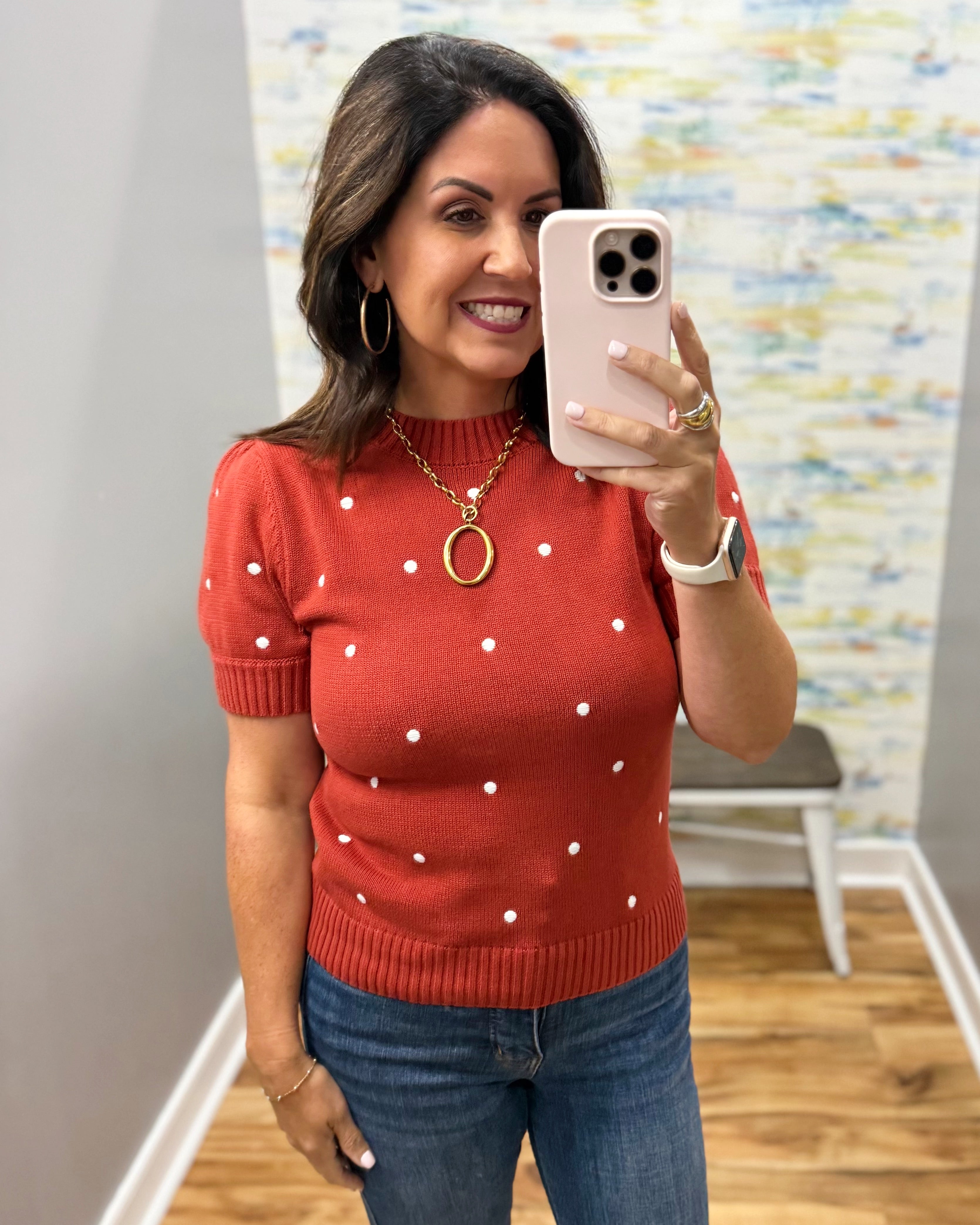 A woman taking a mirror selfie wearing a rust-red short-sleeve knit sweater with white polka dots, paired with blue jeans and gold jewelry.