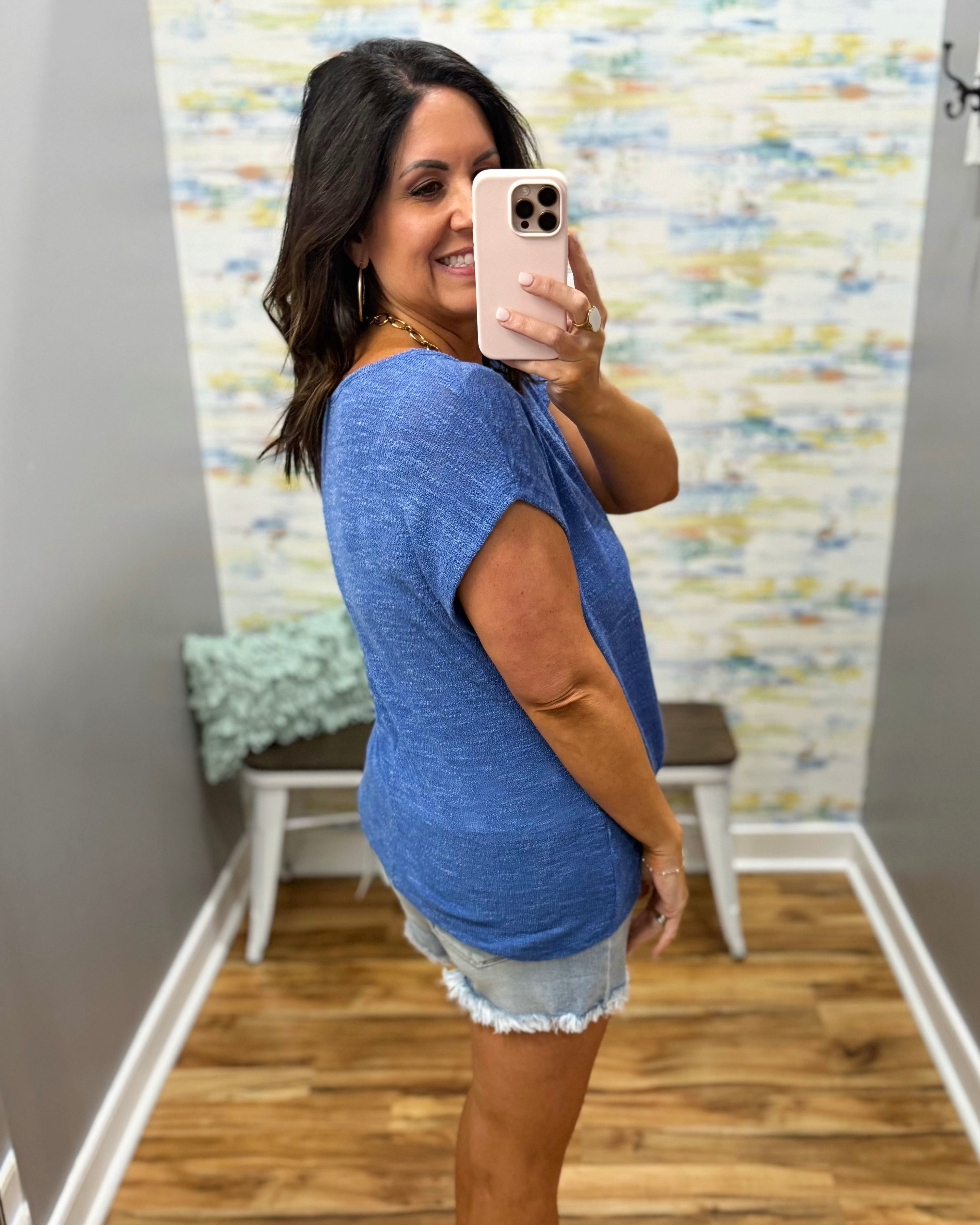 Woman taking a mirror selfie wearing a blue top and denim shorts in a room with a colorful wall.