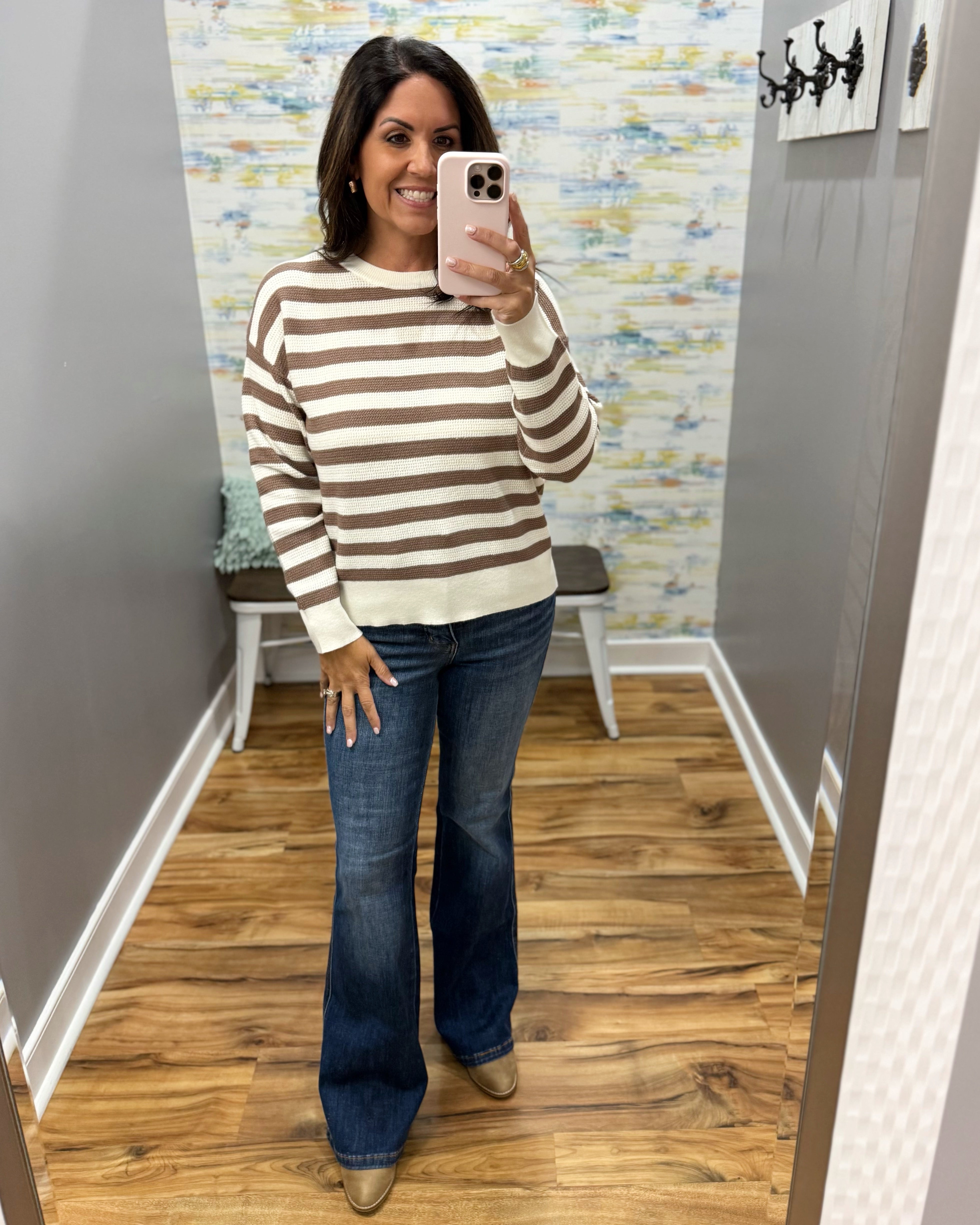 A woman takes a mirror selfie wearing a brown and cream striped long-sleeve sweater, dark blue flare jeans, and tan boots in a home setting with wood floors and gray walls.