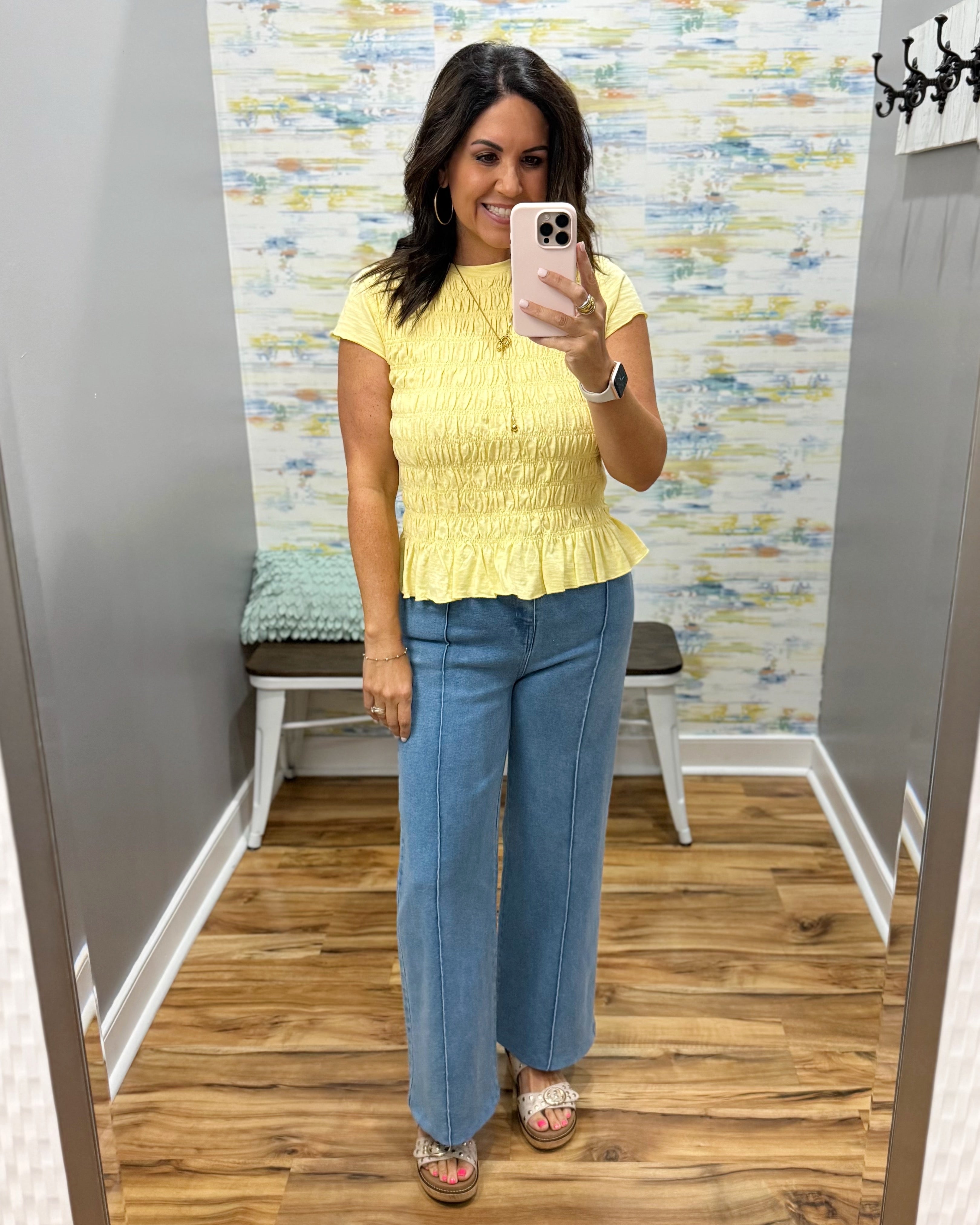 A woman takes a mirror selfie in a fitting room wearing a yellow short-sleeve peplum top and wide-leg blue jeans.