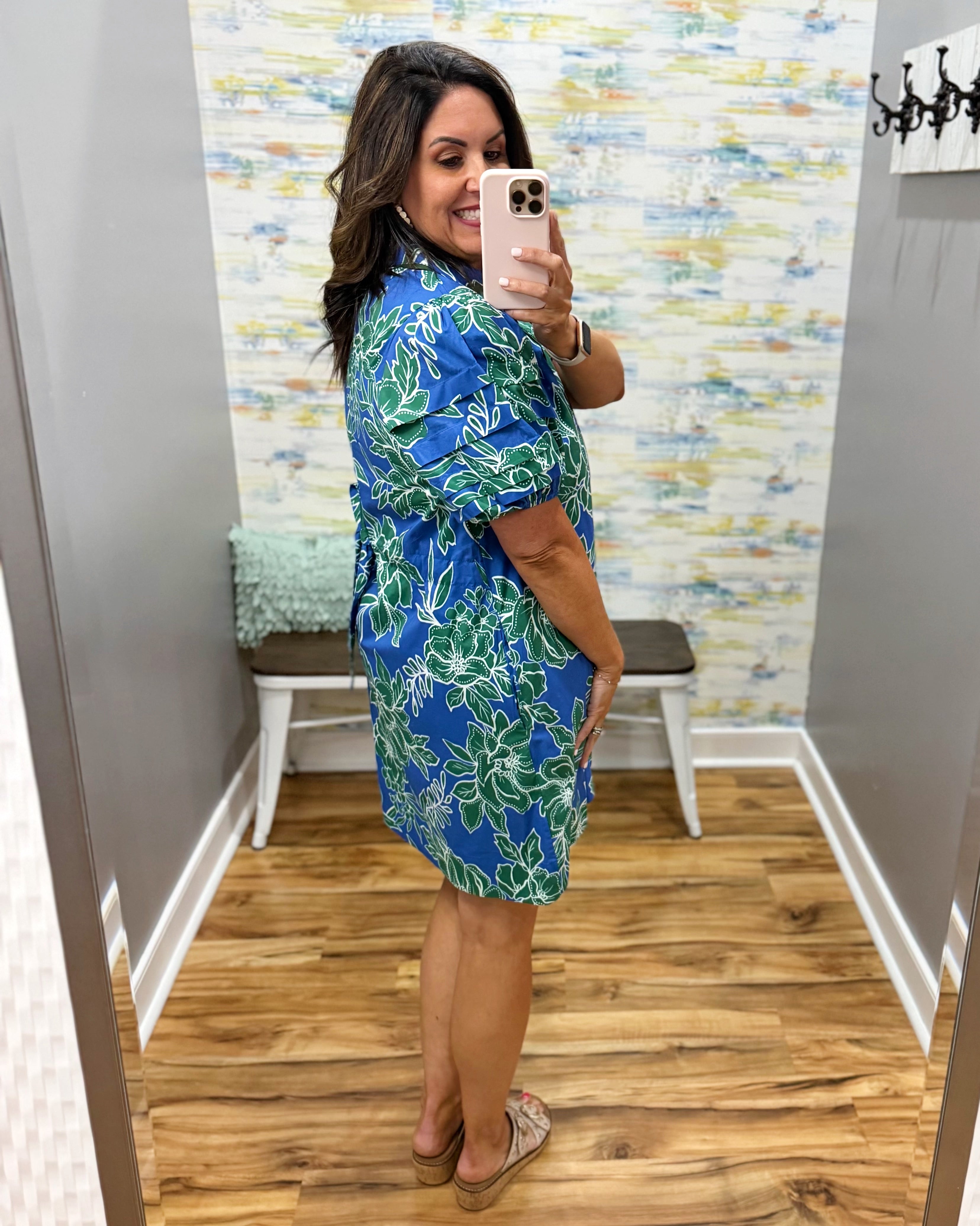 Woman wearing a blue floral dress taking a mirror selfie in a room with a colorful wall.