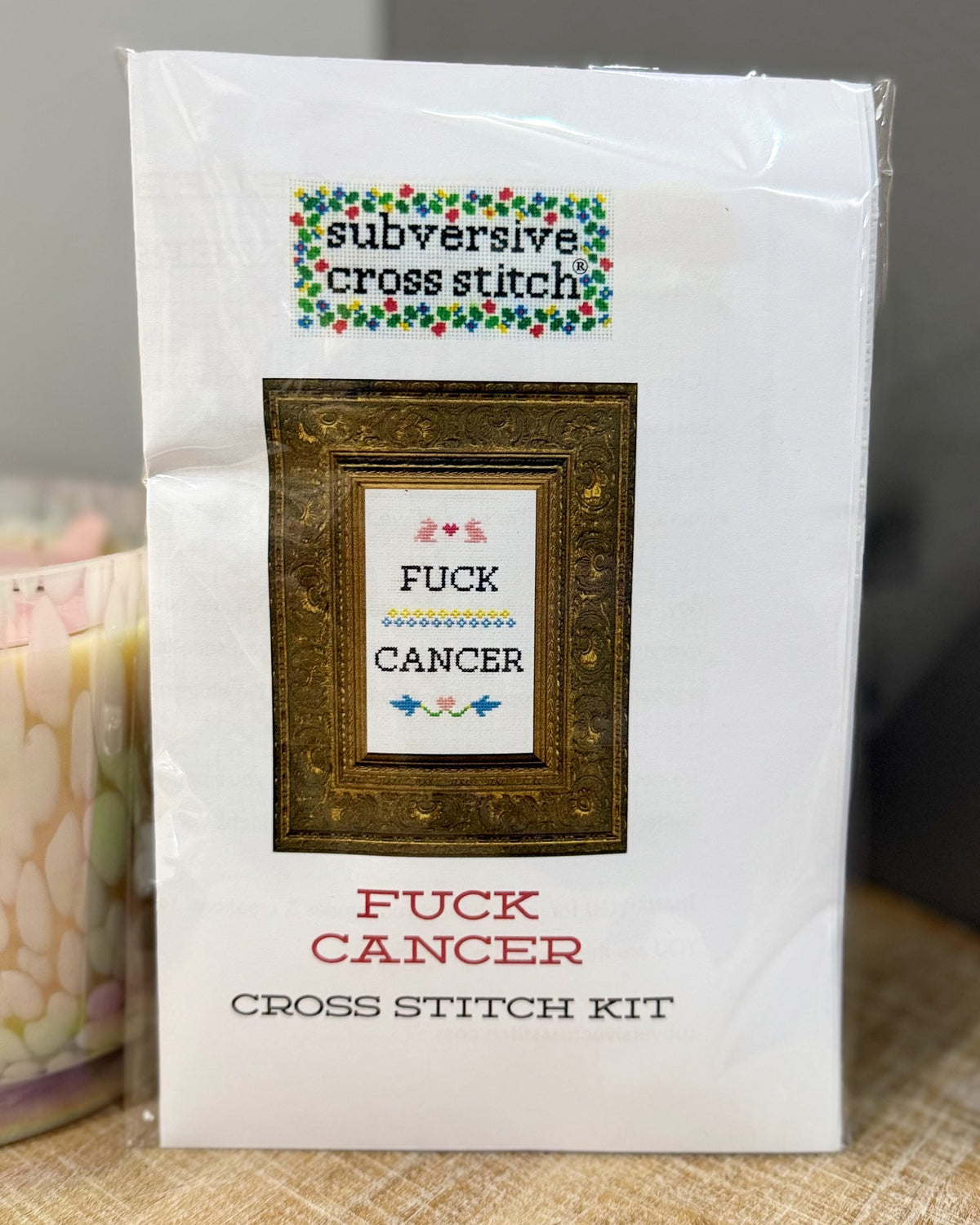 A packaged cross stitch kit labeled 'subversive cross stitch' with the design text 'FUCK CANCER' inside a framed preview, and 'FUCK CANCER CROSS STITCH KIT' printed below on a white package.