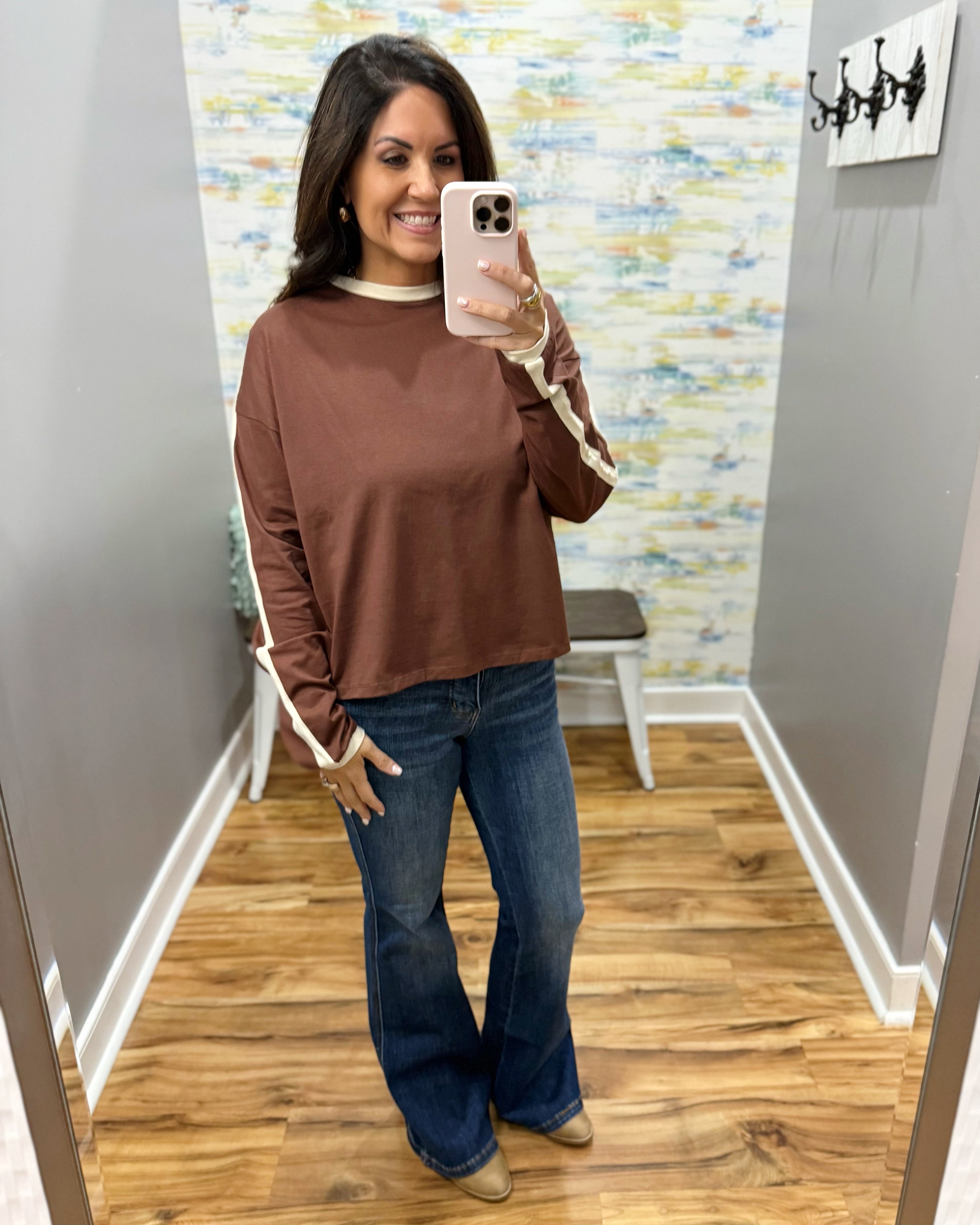 A woman takes a mirror selfie in a fitting room wearing a brown long-sleeve top with beige sleeve stripes, blue jeans, and tan shoes.