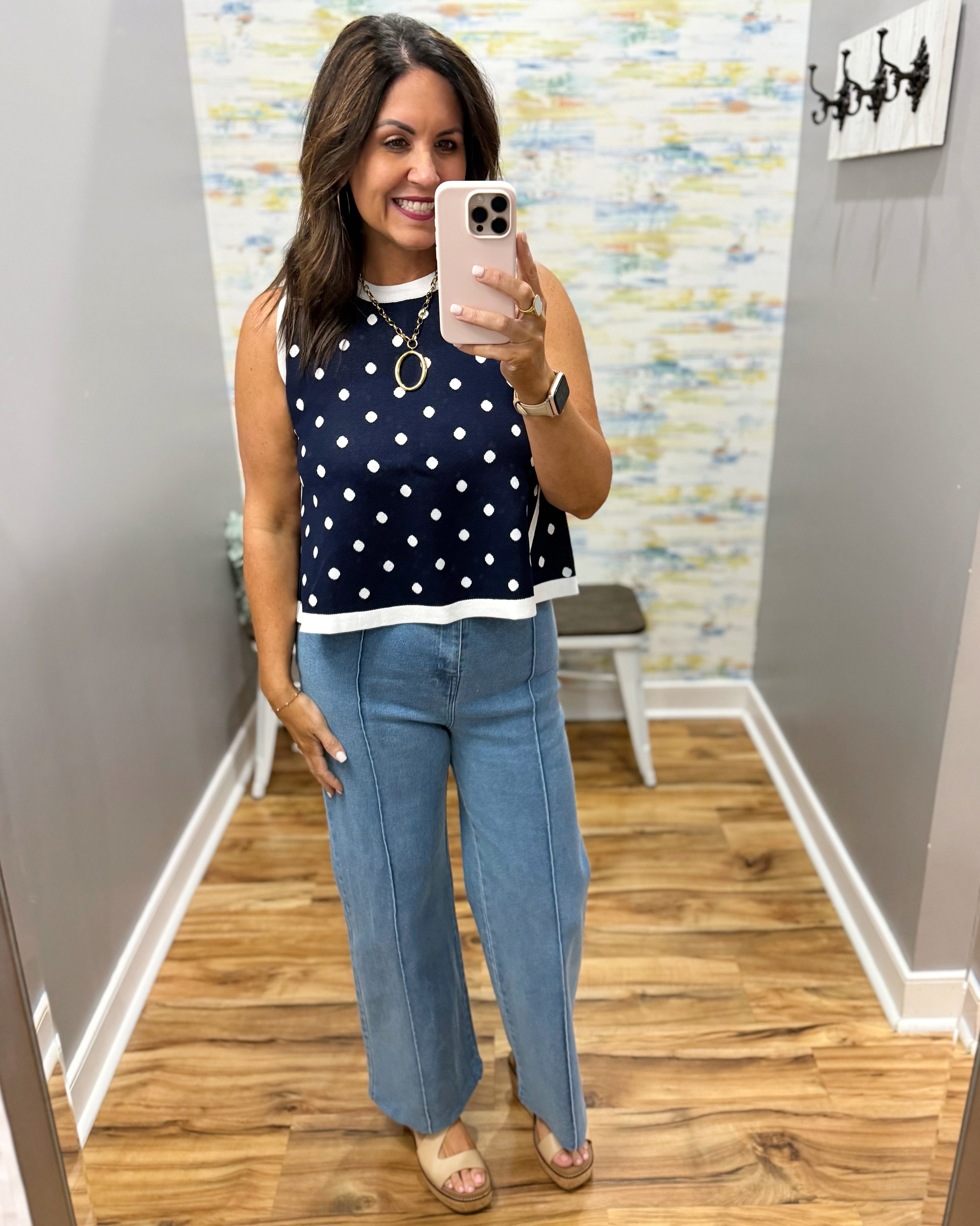 Woman taking a mirror selfie wearing a navy polka dot top and blue jeans in a room with a textured wall.