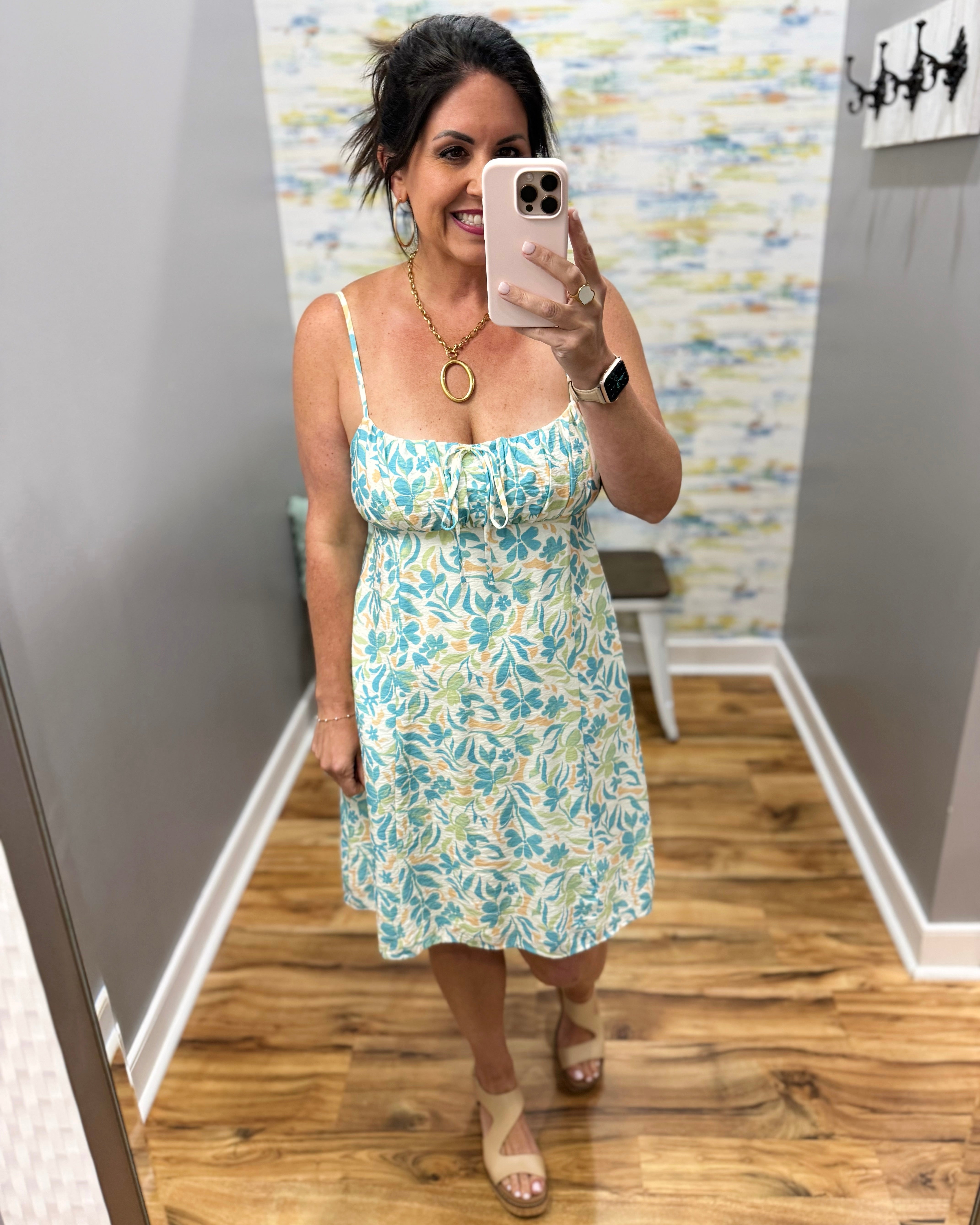 Woman taking a mirror selfie wearing a floral dress in a room with wooden flooring and a patterned wall.