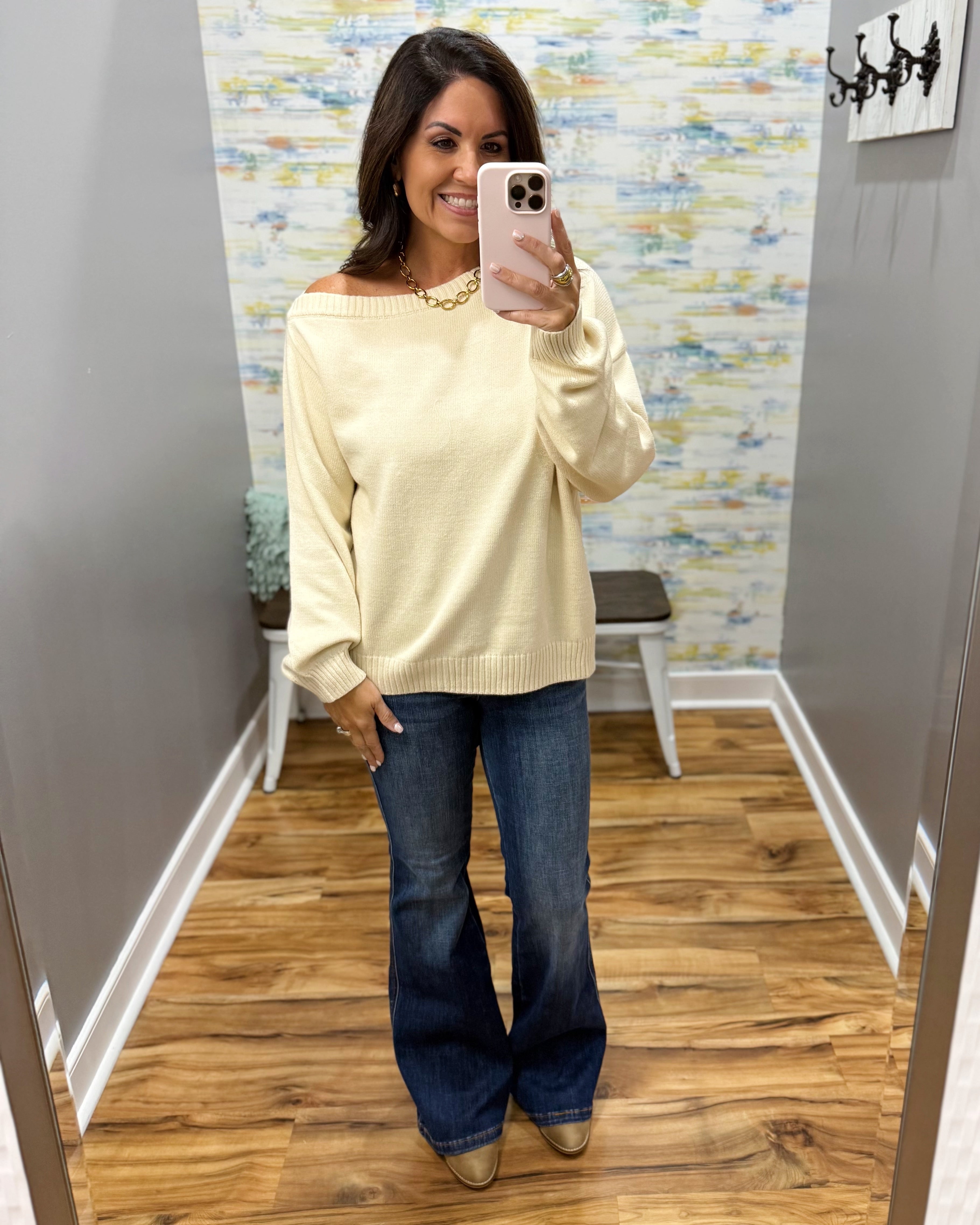A woman takes a mirror selfie in a fitting room wearing a pale yellow off-the-shoulder knit sweater, blue flare jeans, and tan pointed shoes.