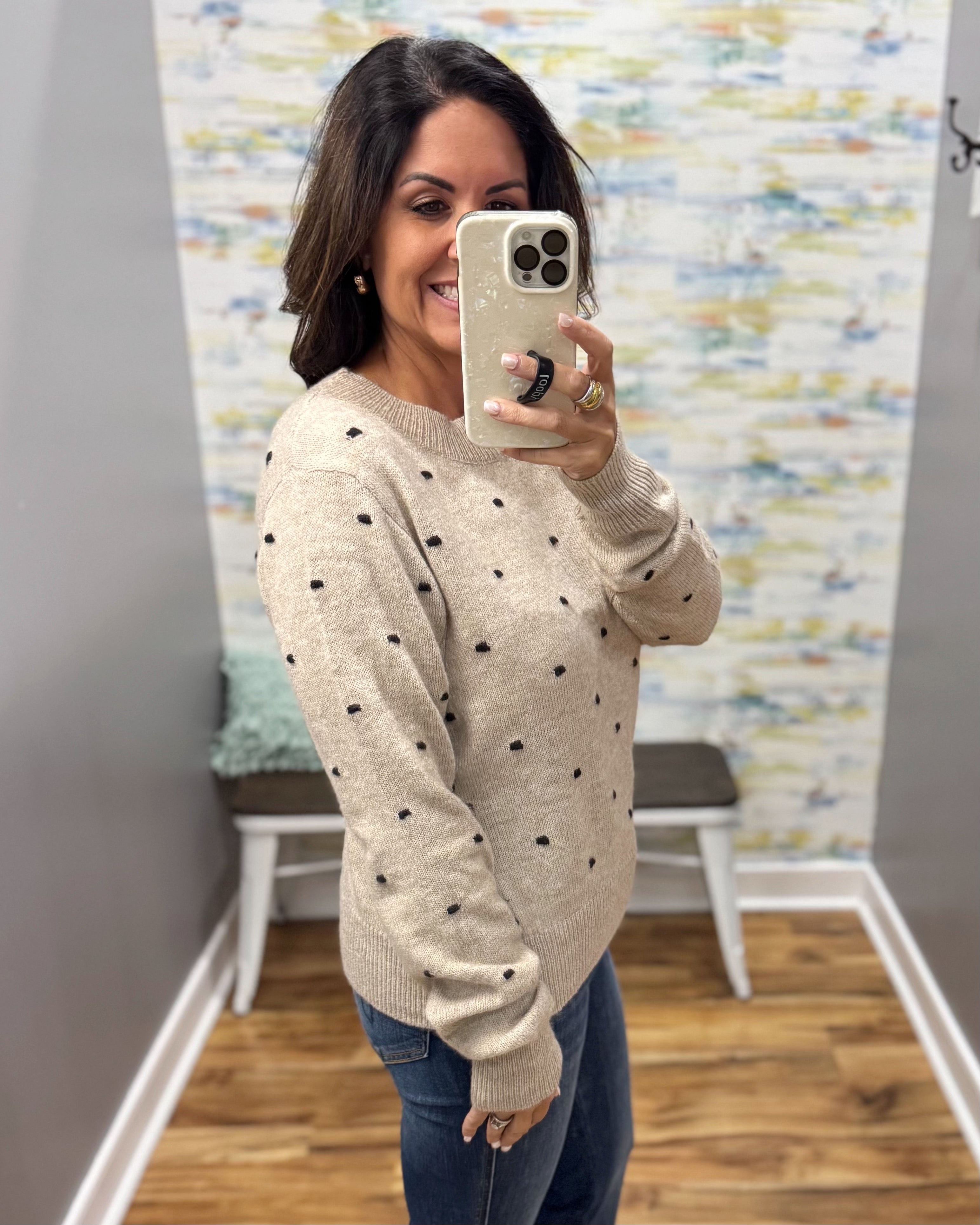A woman takes a mirror selfie wearing a beige knit sweater with small black polka dots and blue jeans, standing in a room with a patterned wallpaper background.