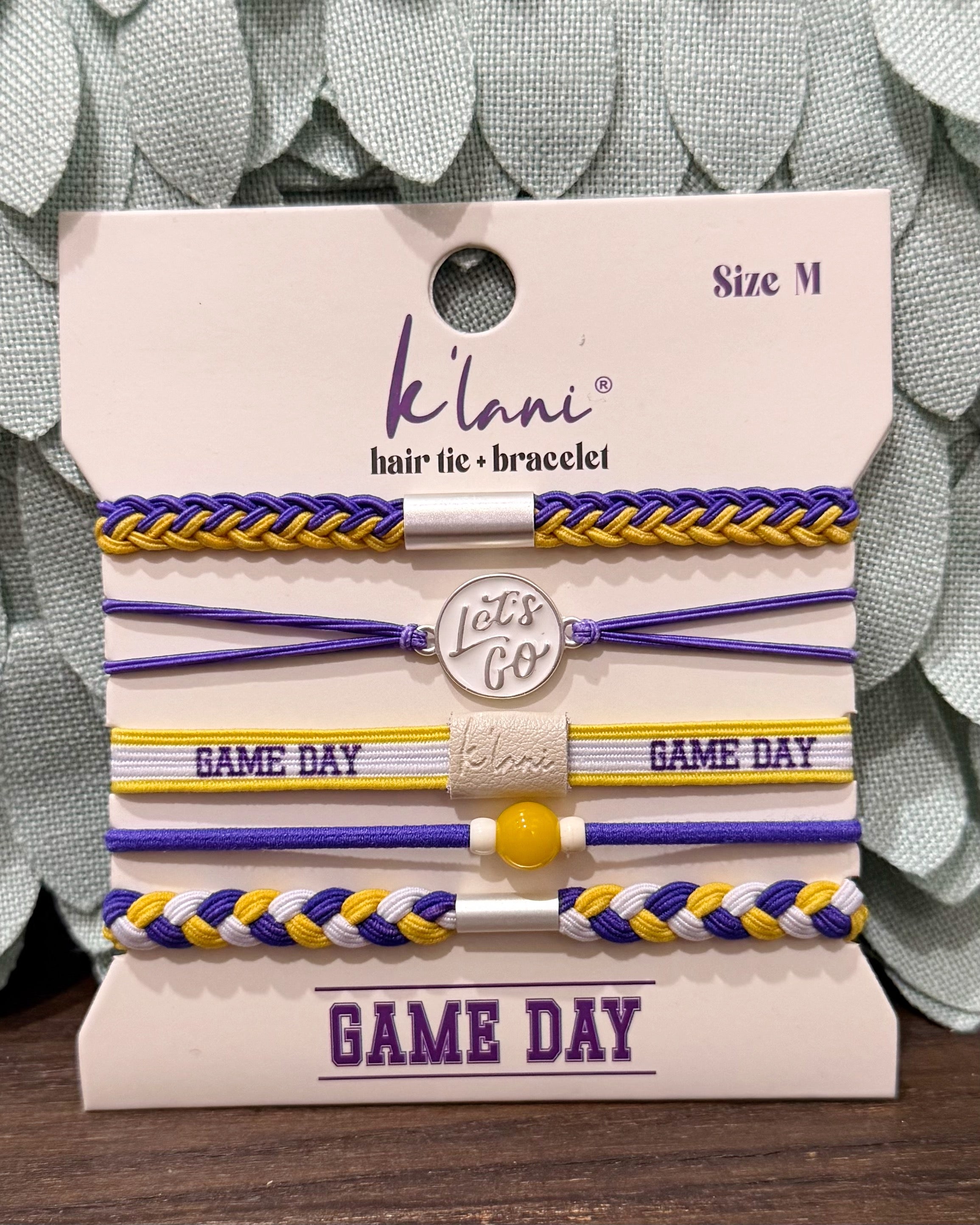 A set of five K'lan's hair tie bracelets in purple, yellow, and white, displayed on a branded card. The set includes braided and flat elastic bands with 'GAME DAY' text and a central charm that reads 'Let's Go'.