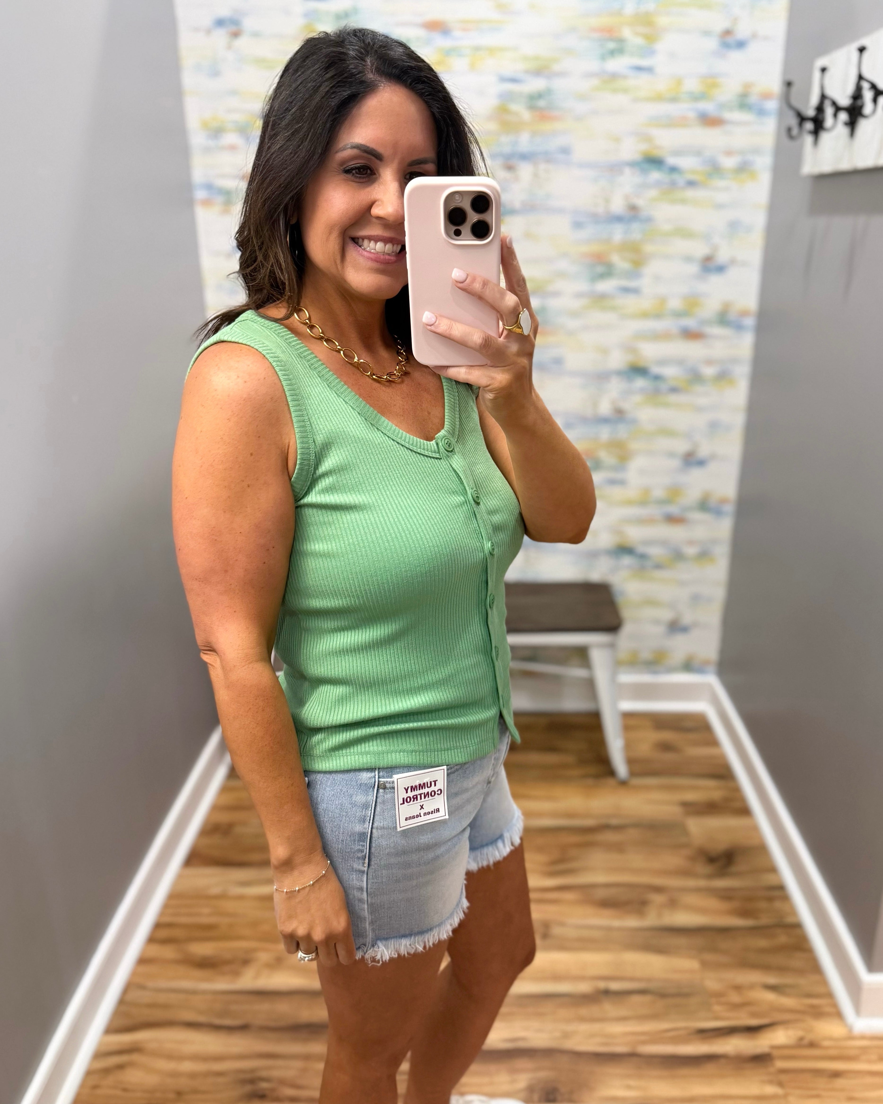 Woman taking a mirror selfie wearing a green tank top and denim shorts in a room with a colorful wall.