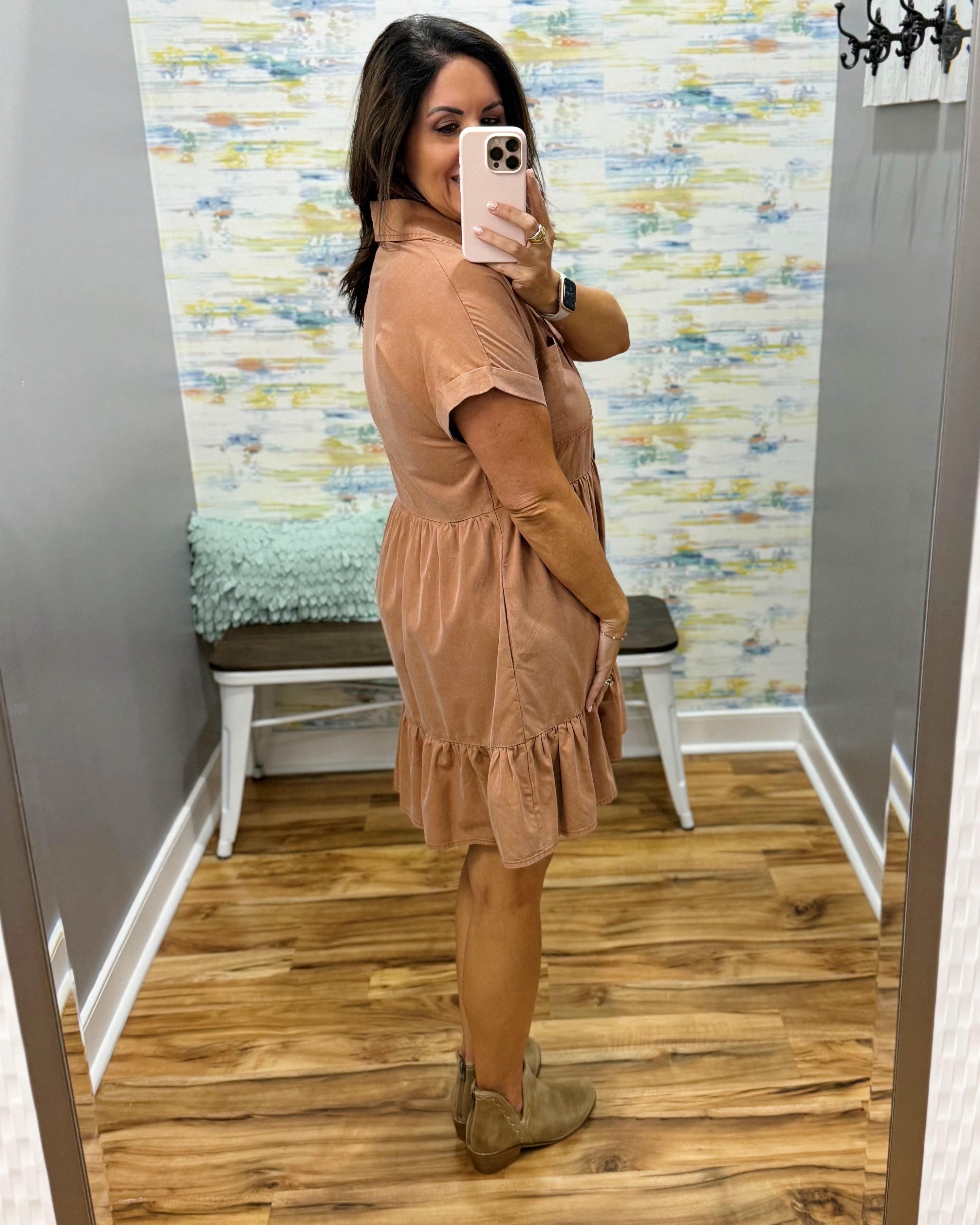 A woman takes a mirror selfie in a fitting room wearing a short-sleeve tan dress with a ruffled hem and tan ankle boots.