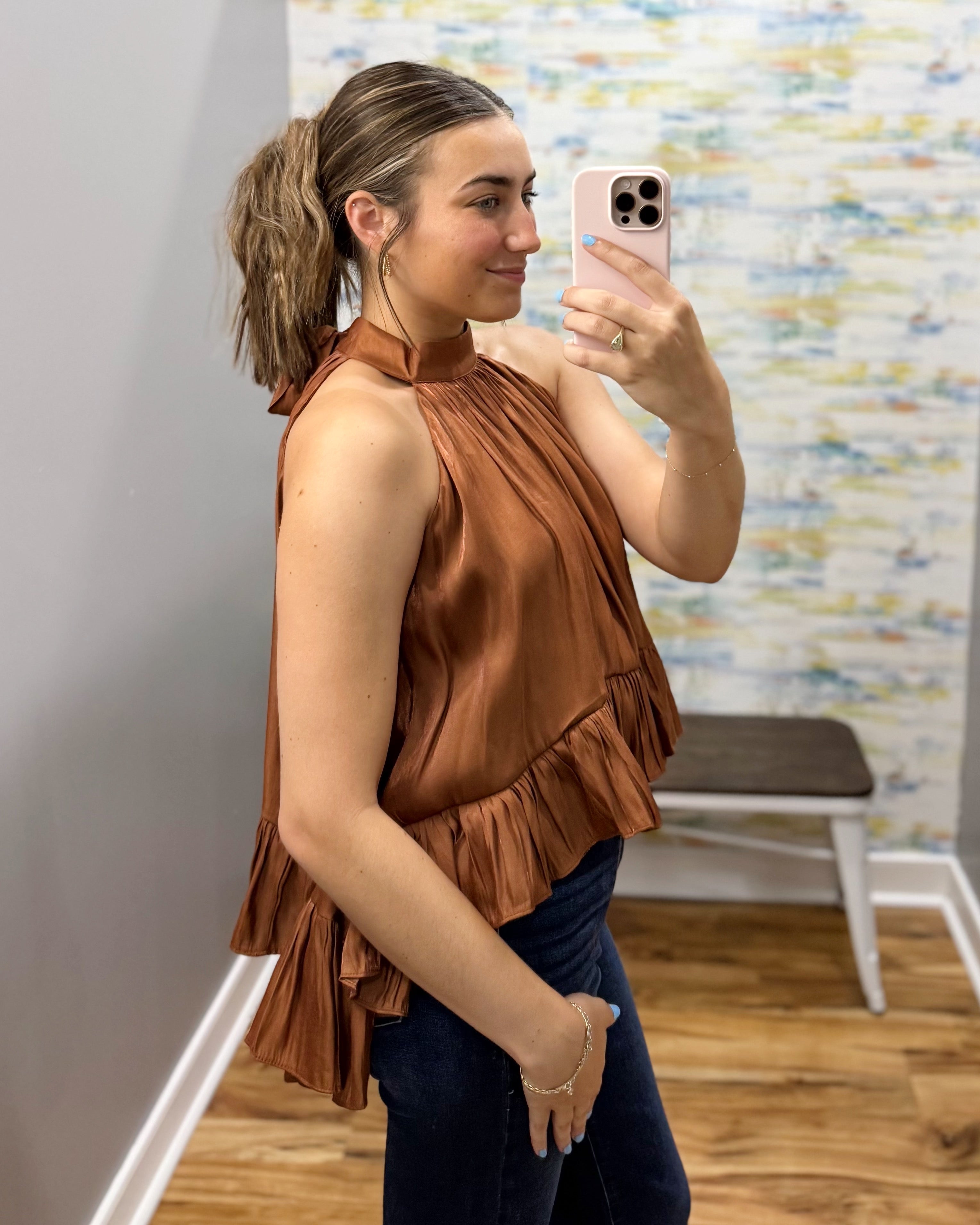 A woman takes a mirror selfie wearing a sleeveless, high-neck, ruffled bronze-brown top and dark blue jeans in a fitting room with a patterned wall and wooden floor.
