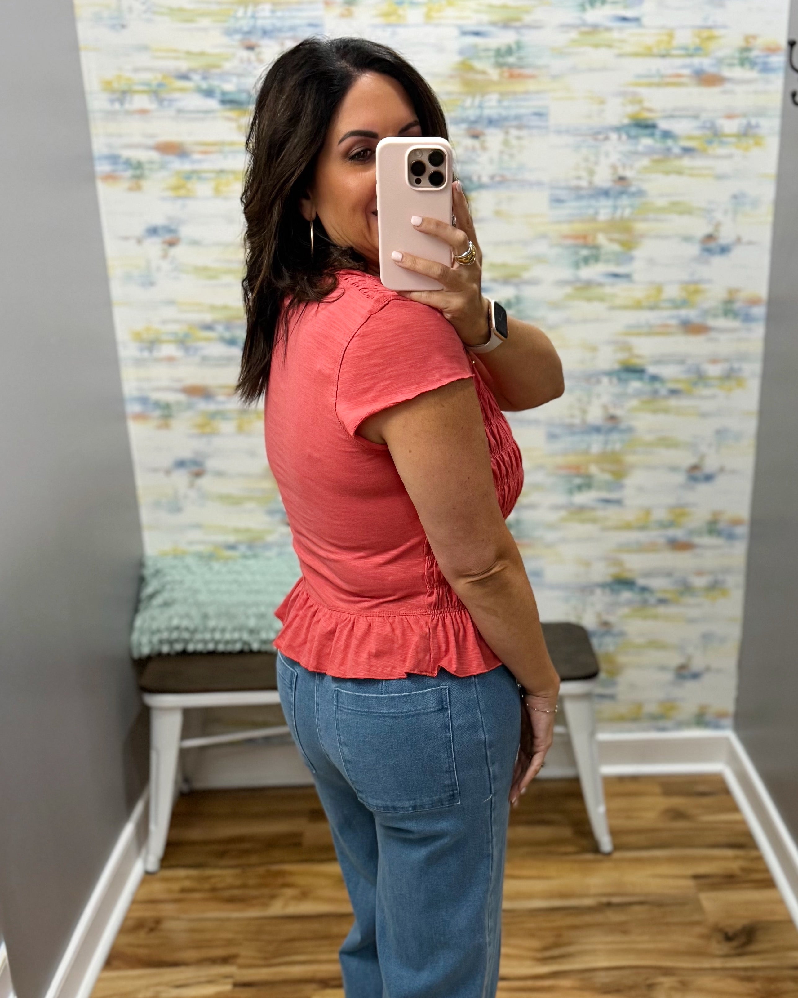 A woman takes a mirror selfie in a fitting room wearing a coral pink short-sleeve ruffled top and blue jeans.