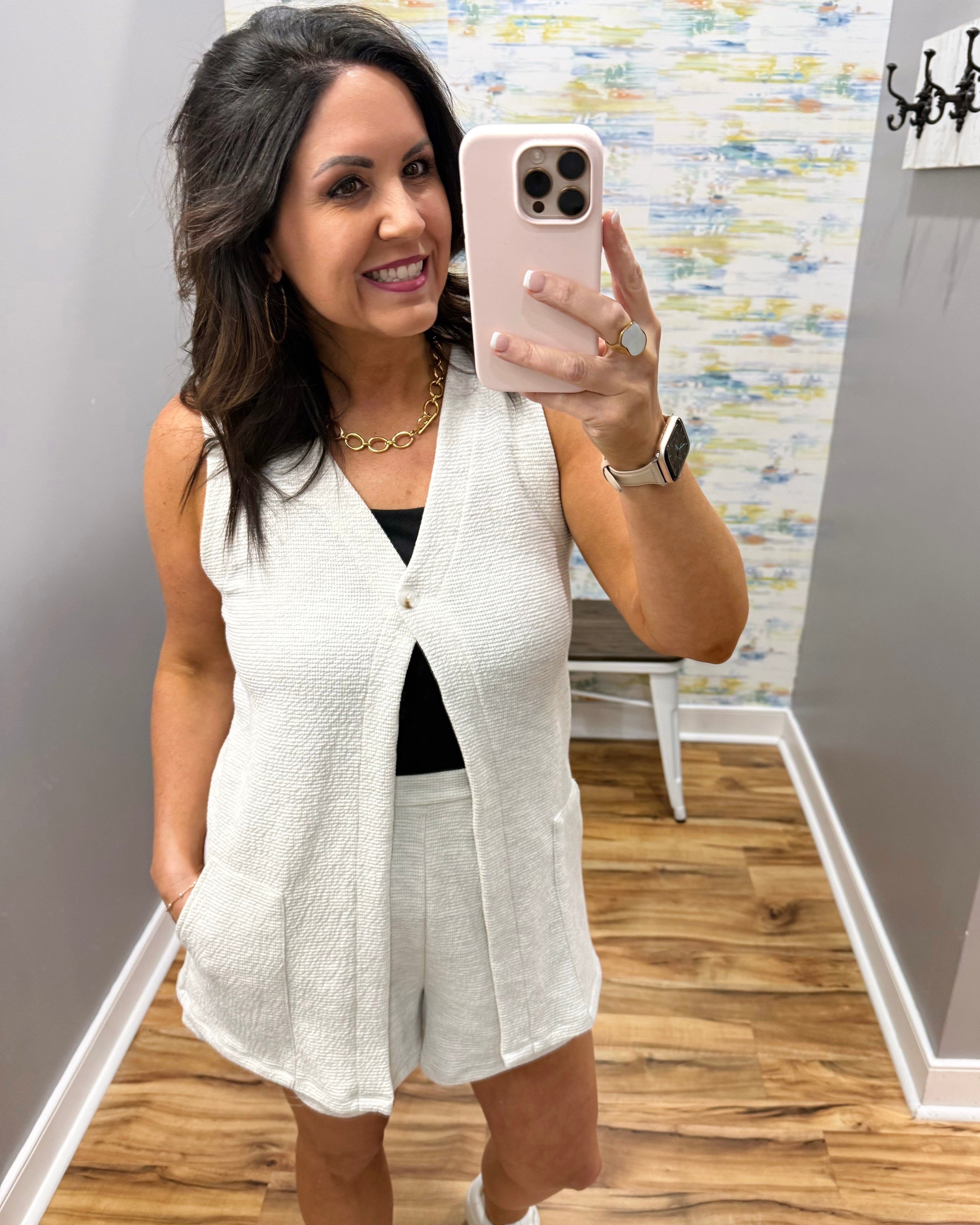 Woman taking a mirror selfie wearing a white vest and shorts in a room with a colorful wall.
