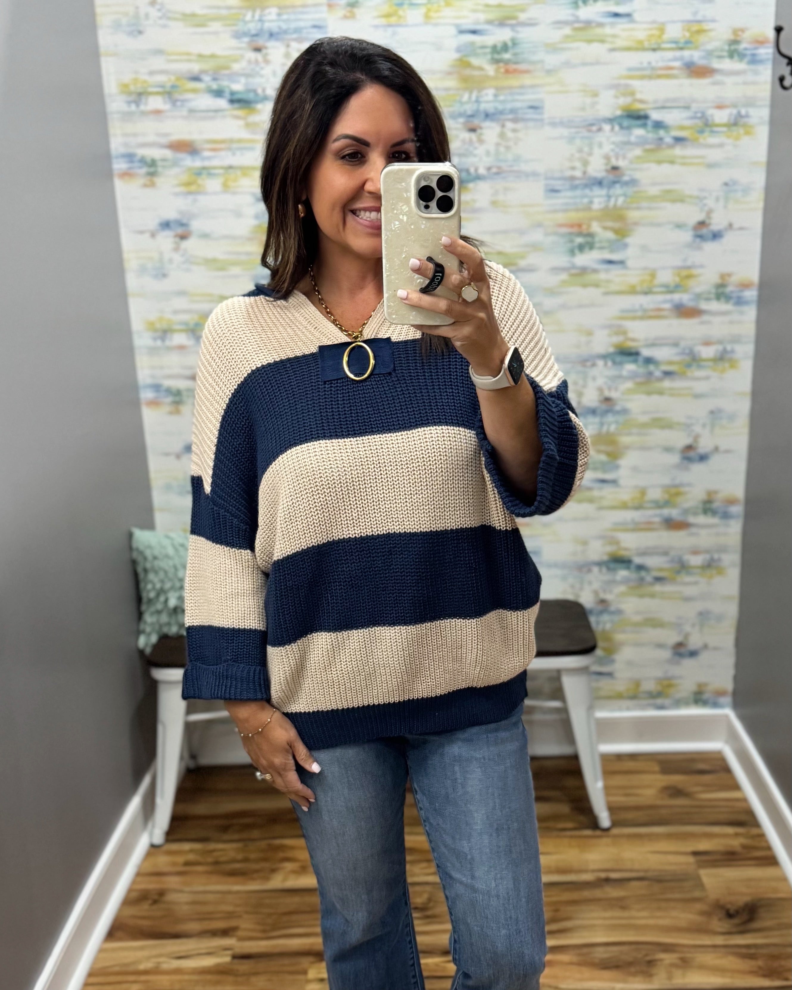 A woman takes a mirror selfie in a boutique showing a knit sweater with horizontal navy blue and cream stripes, paired with blue jeans.