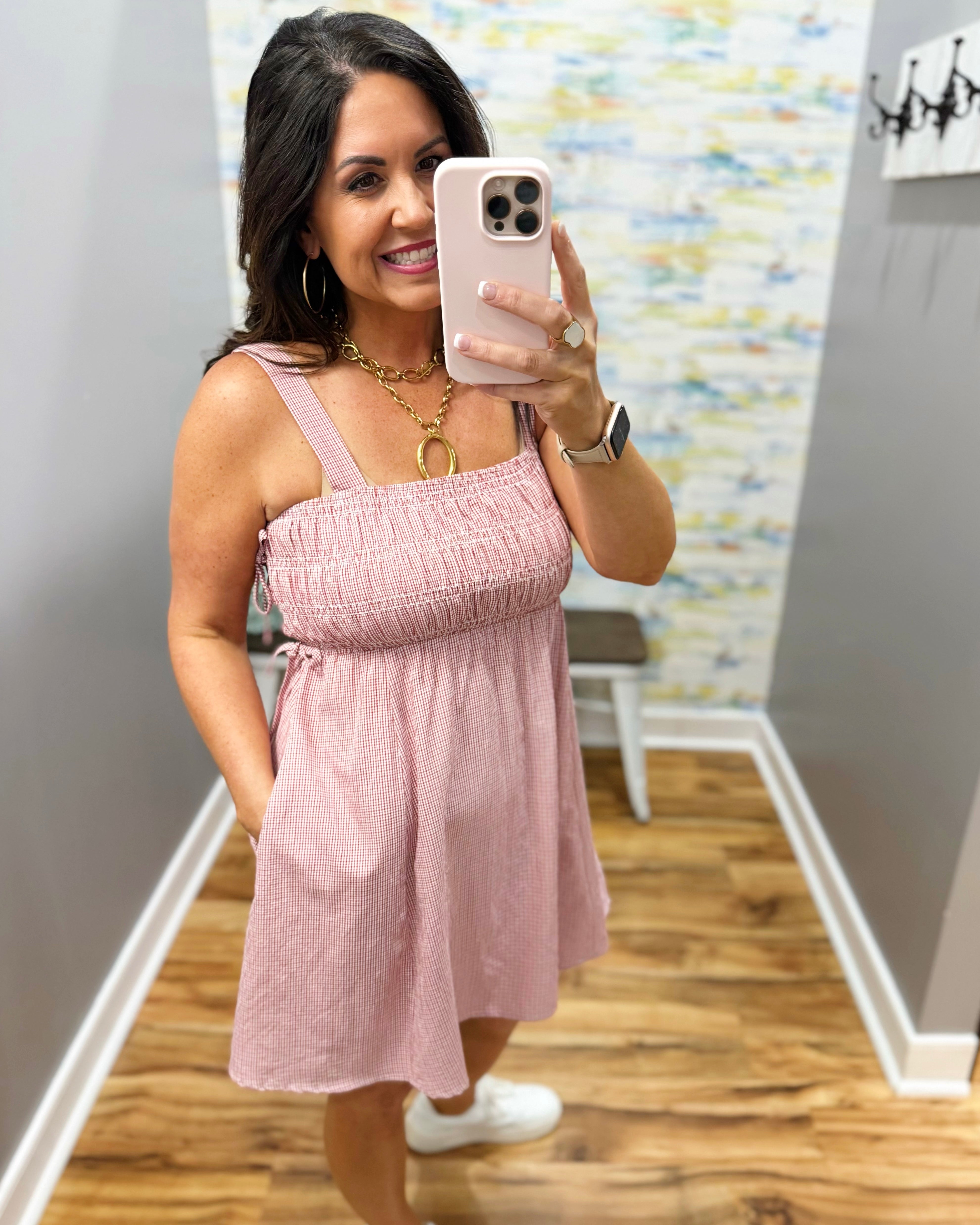 Woman taking a mirror selfie wearing a pink dress in a room with colorful wall art.